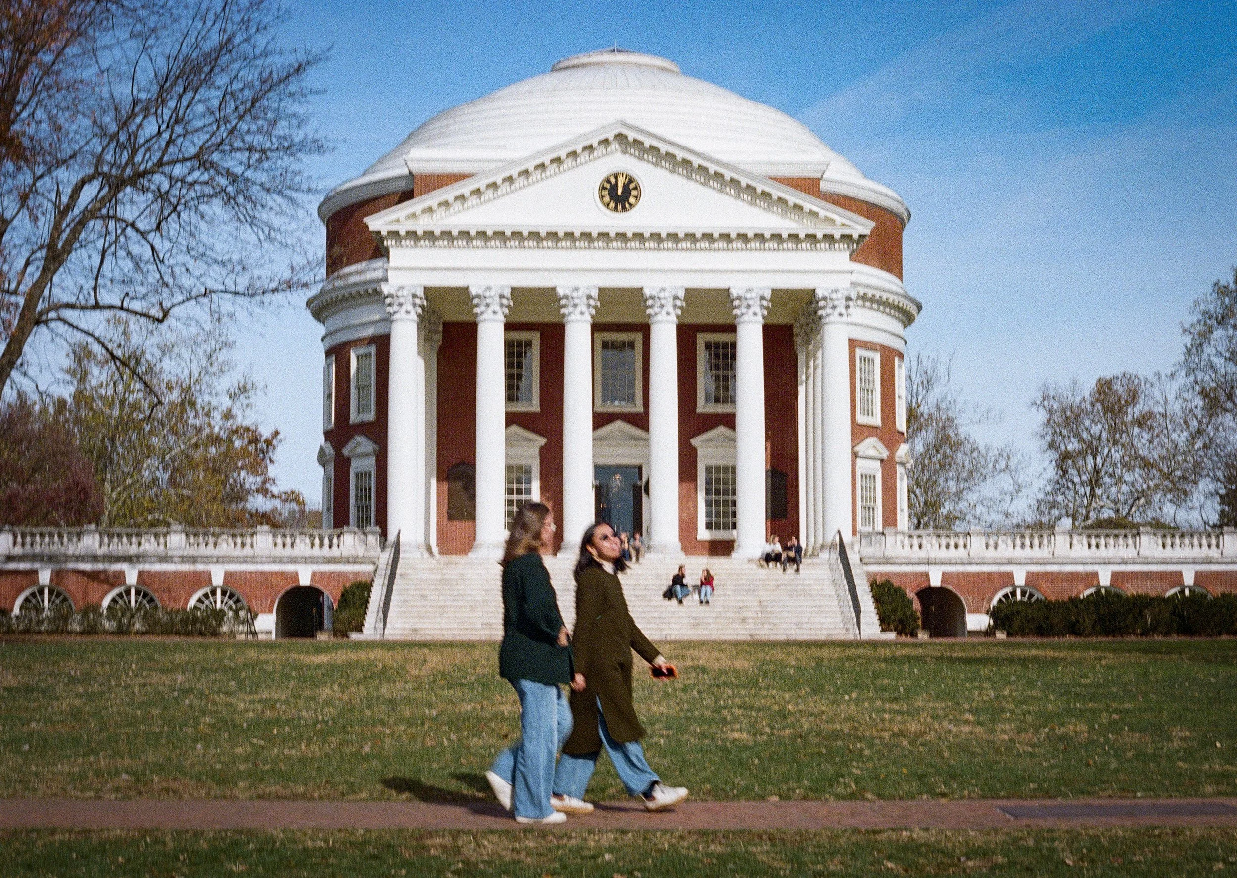 People walking on a sidewalk in front of a large, historic red brick building with white columns, a clock, and a domed roof, trees on either side, blue sky overhead.