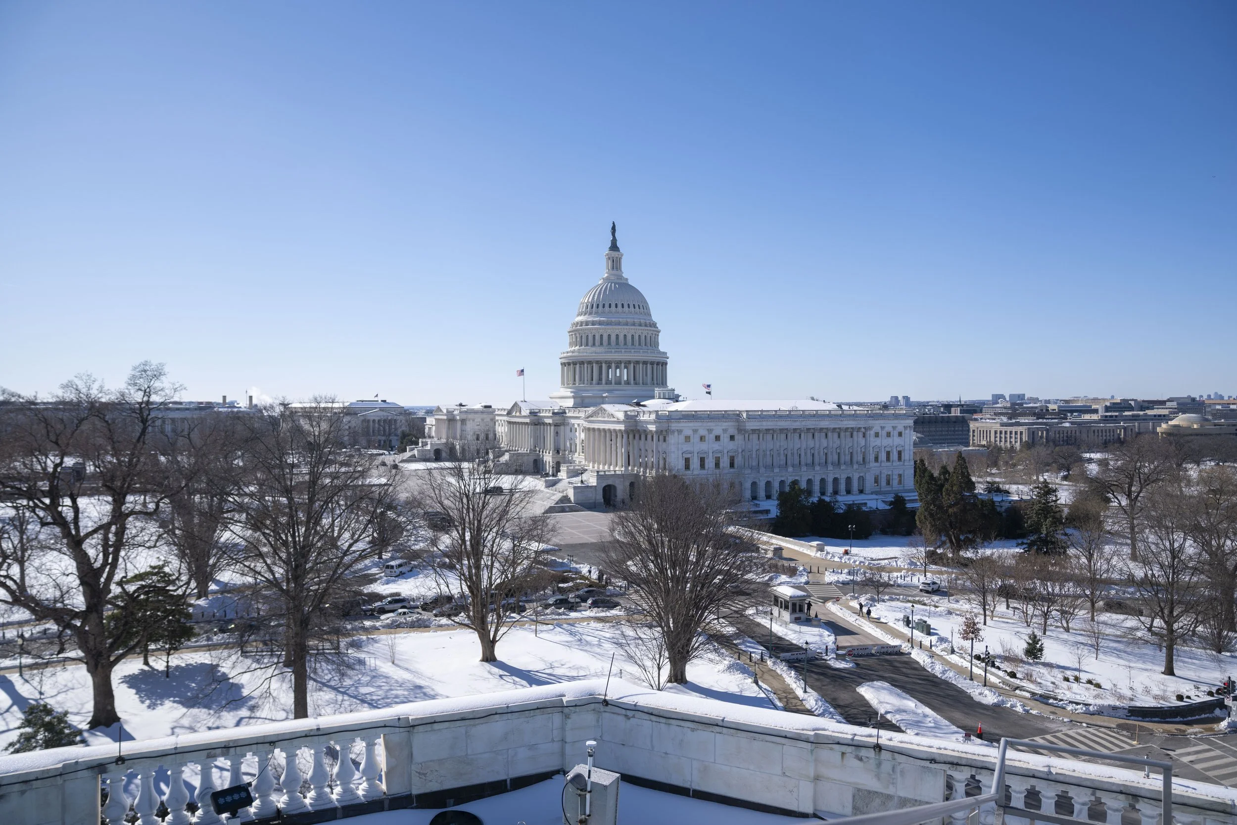 Snow-covered grounds and trees surround the United States Capitol building in Washington D.C., with the sky clear and blue.