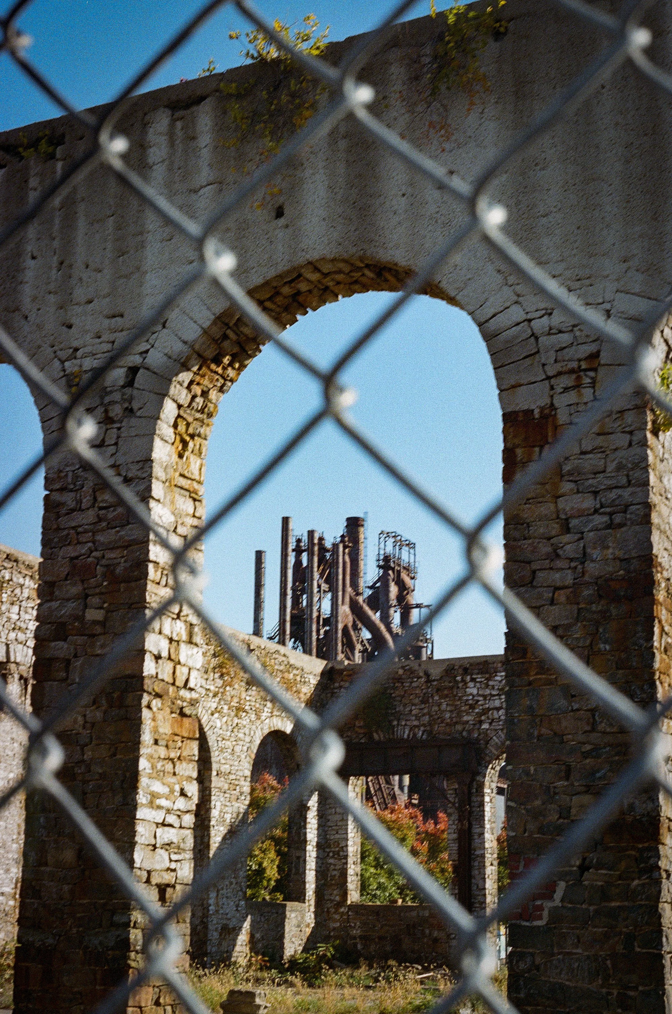 View of stone arches and structure seen through a chain-link fence at an abandoned or ruined building with blue sky overhead.