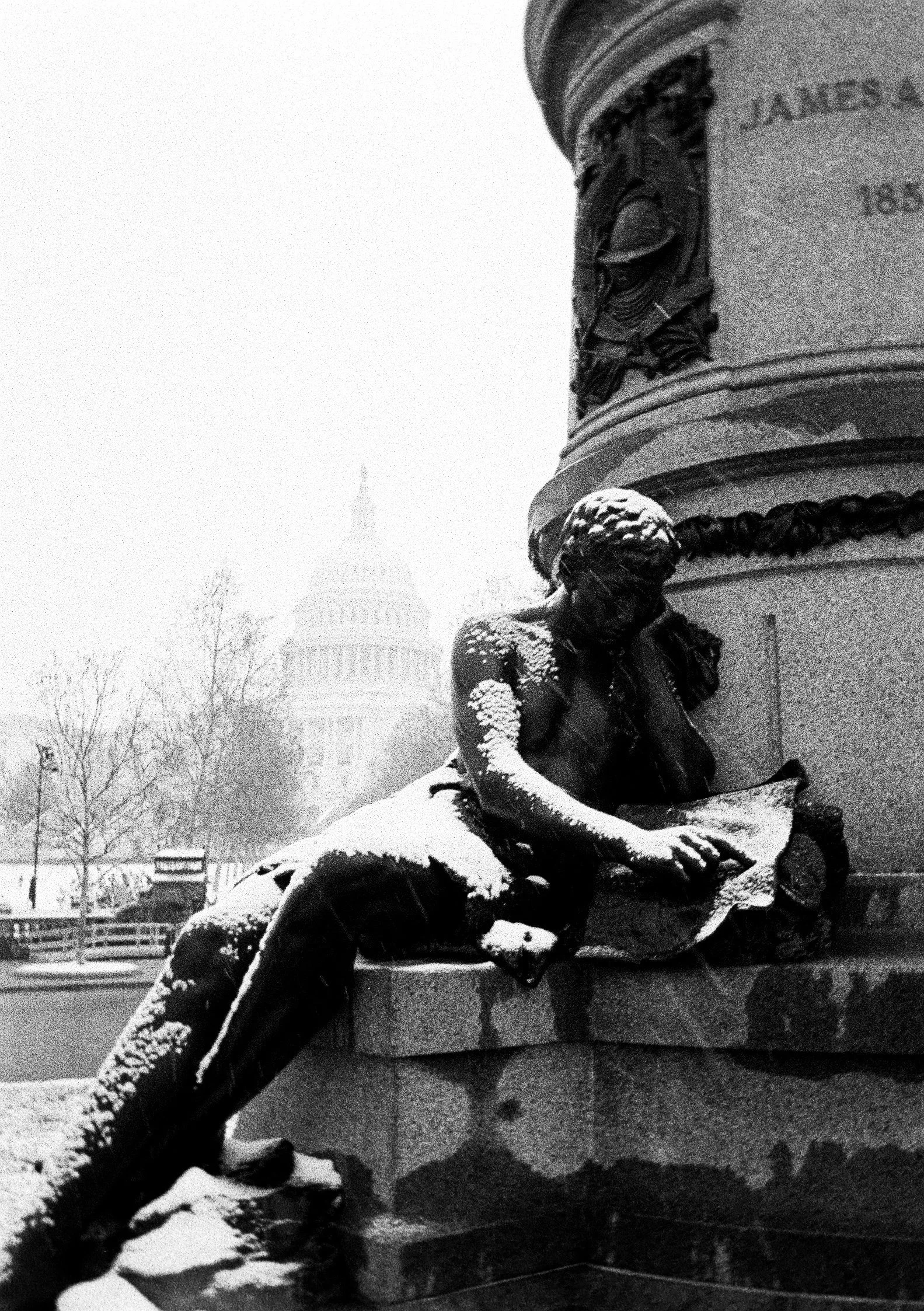 Snow-covered statue of a seated woman holding an open book at night, with the U.S. Capitol building visible in the background.