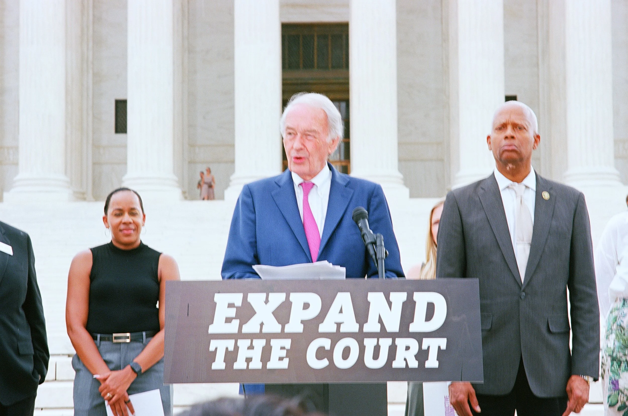 Sen. Ed Markey, wearing a blue suit and pink tie, is speaking at a podium with a sign that reads 'EXPAND THE COURT.' 