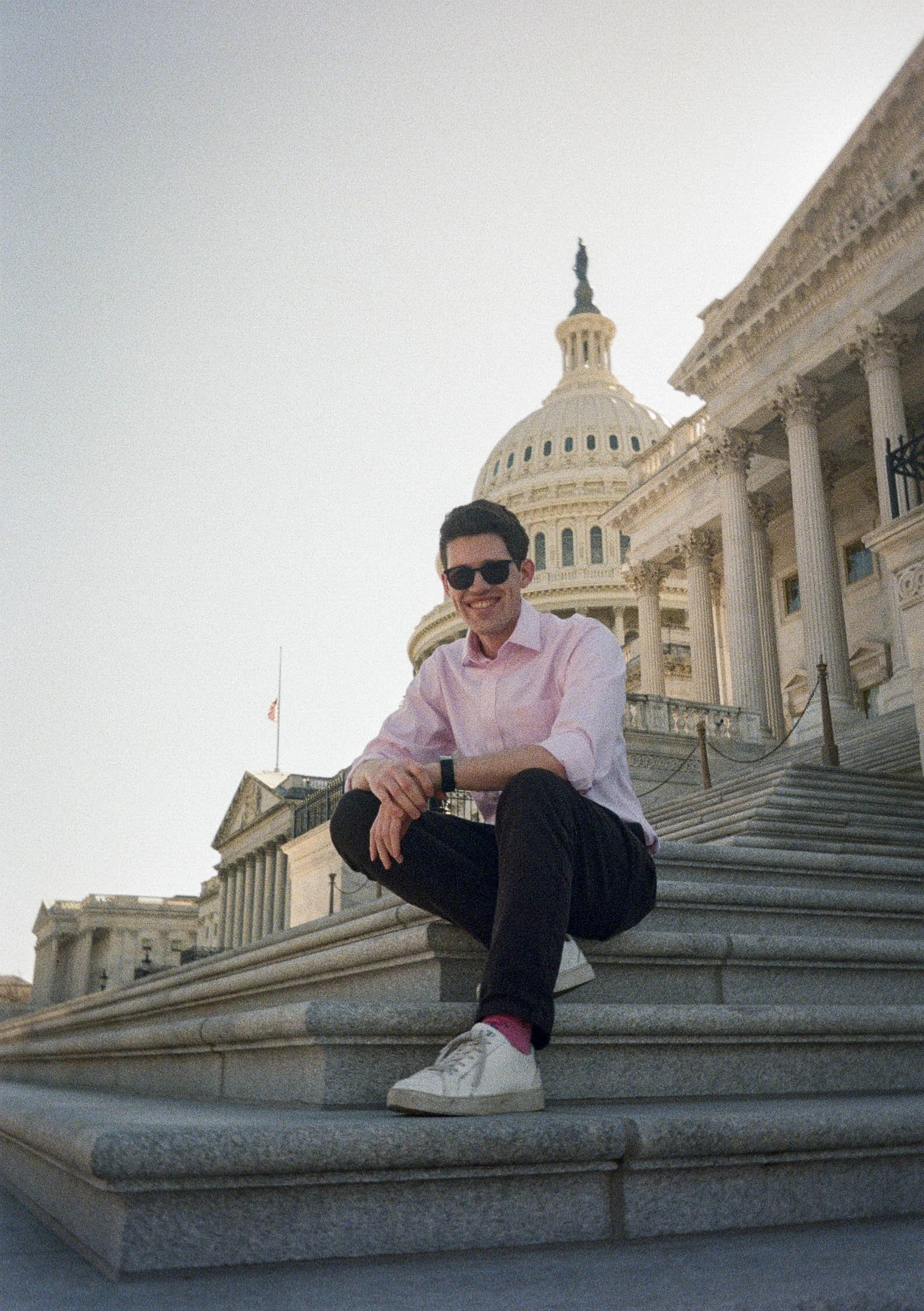 Brooks, wearing a light pink shirt and black pants, sitting on the steps of the United States Capitol building, smiling at the camera during daylight hours.