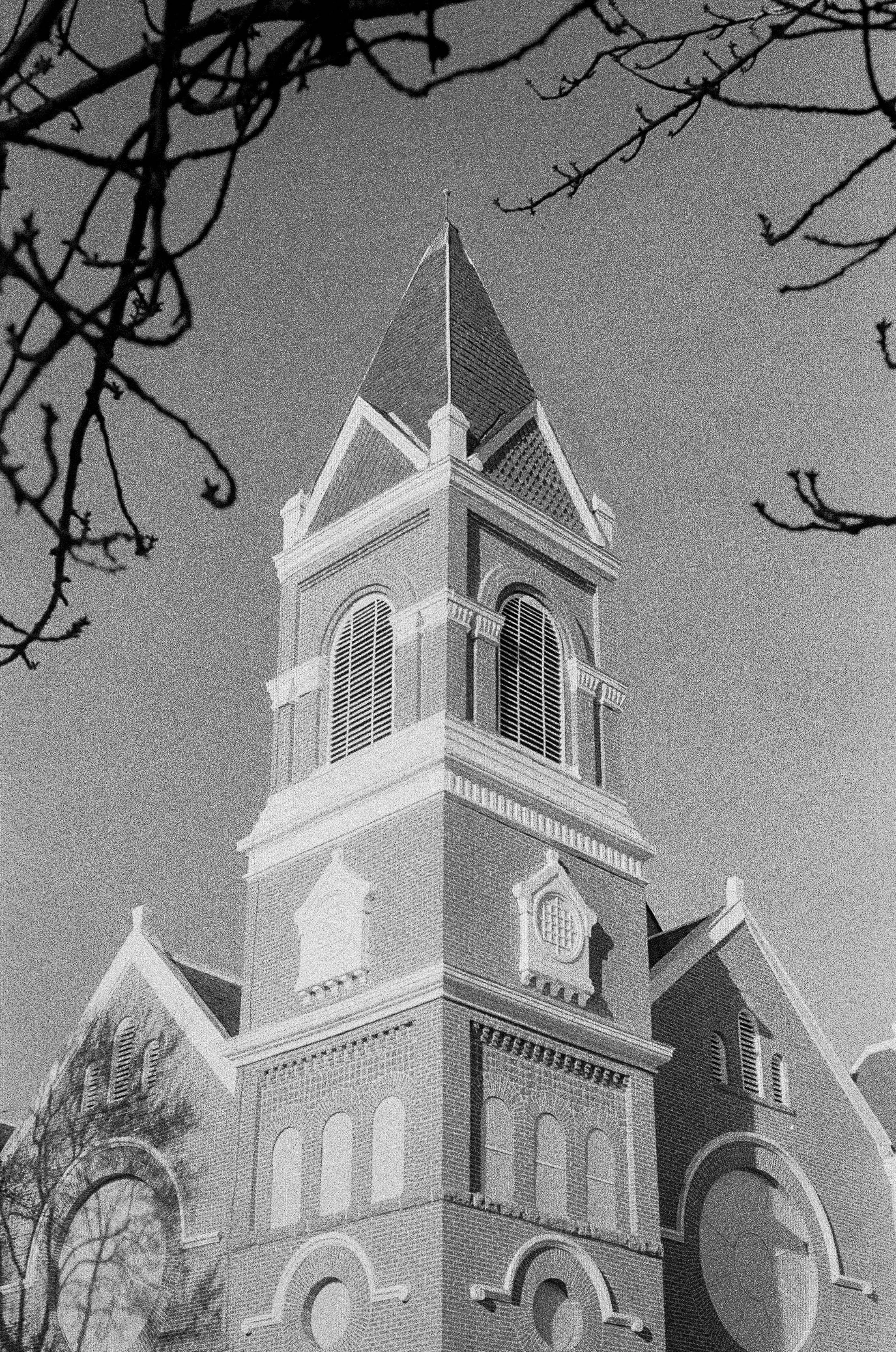 Black and white photo of a church tower with a pointed roof, ornate windows, and decorative architectural details, framed by leafless tree branches.