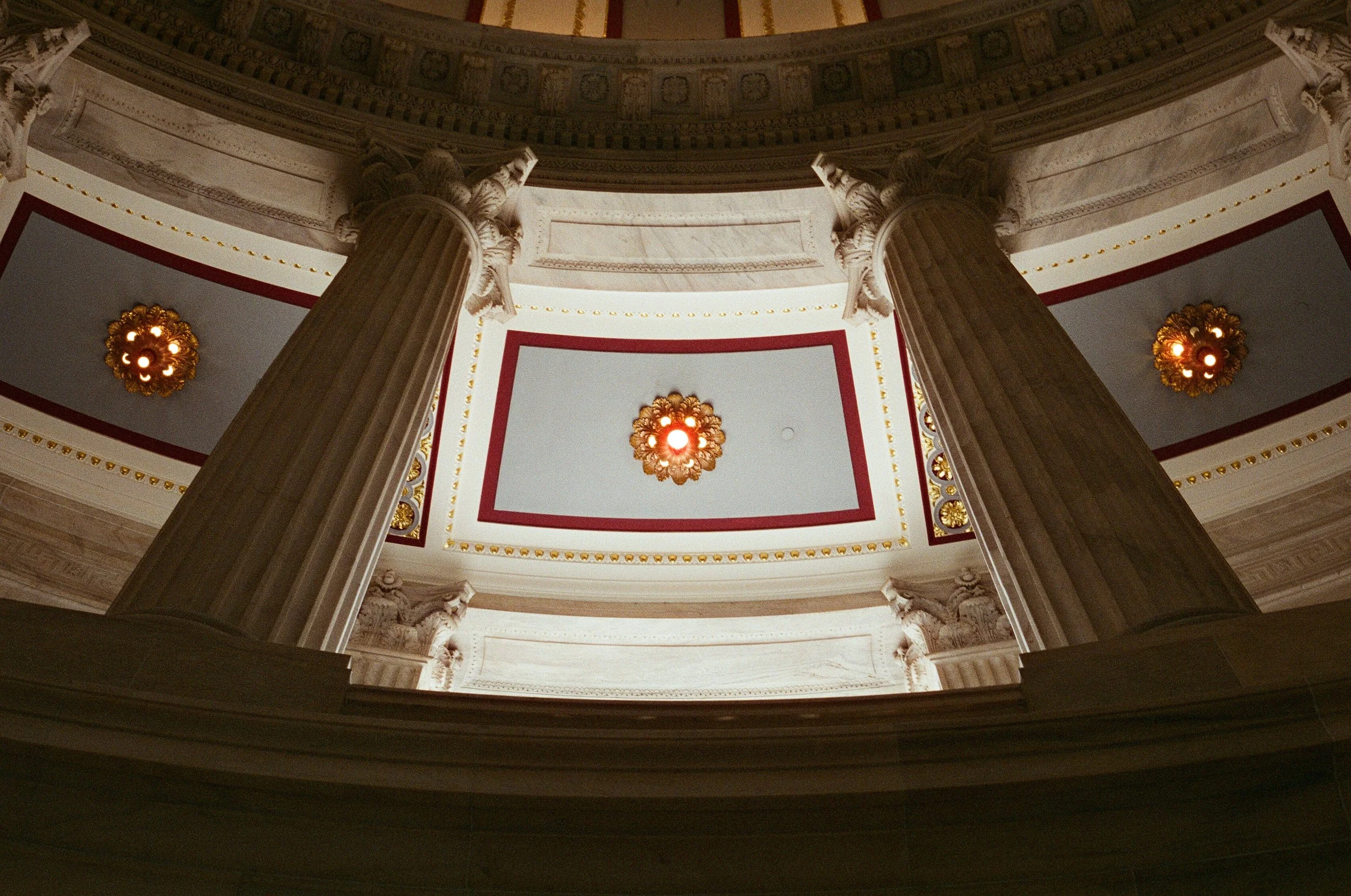 Interior view of a grand building with tall marble columns and ornate ceiling details, viewed from the ground looking up.