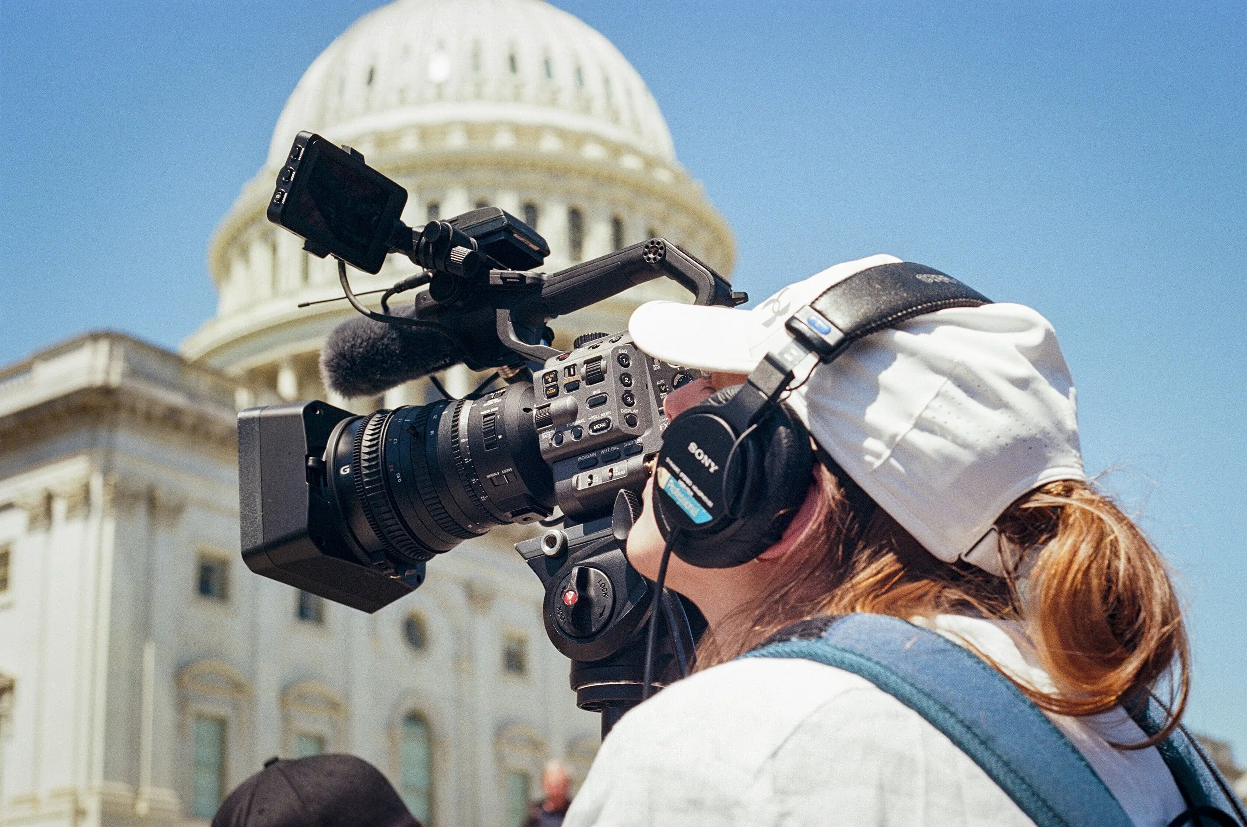 A person with red hair operating a professional video camera outdoors in front of the US Capitol building with a dome, under a clear blue sky.