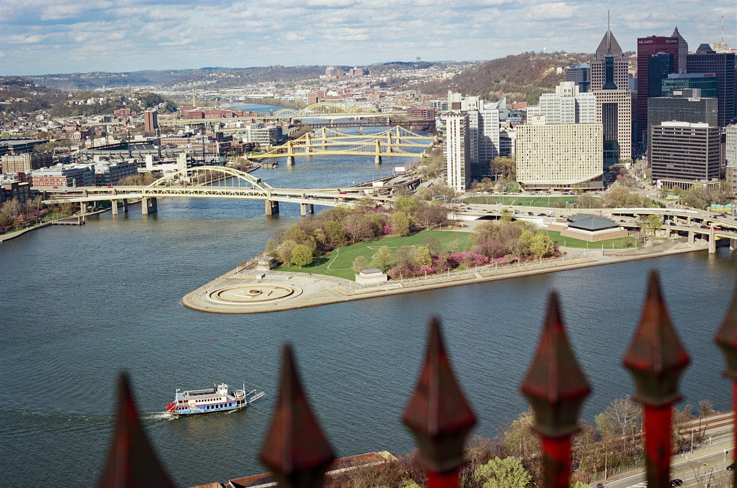 A city skyline with tall buildings and bridges over a river, viewed through an orange metal fence in the foreground.