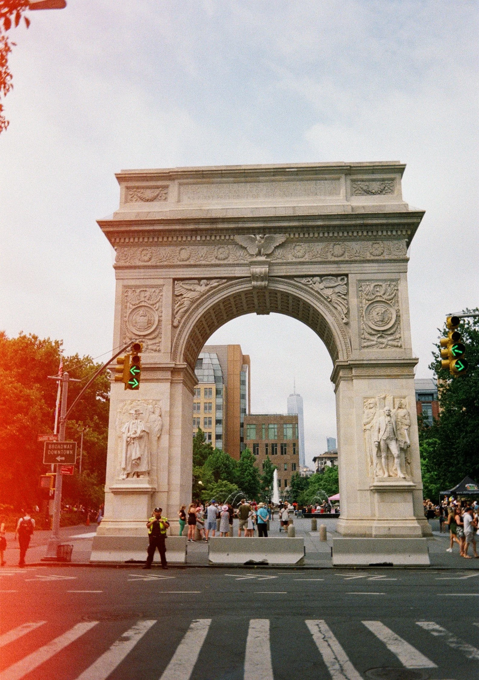 View of the Washington Square Arch in New York City with people walking and tourists gathered underneath it, trees and buildings in the background.