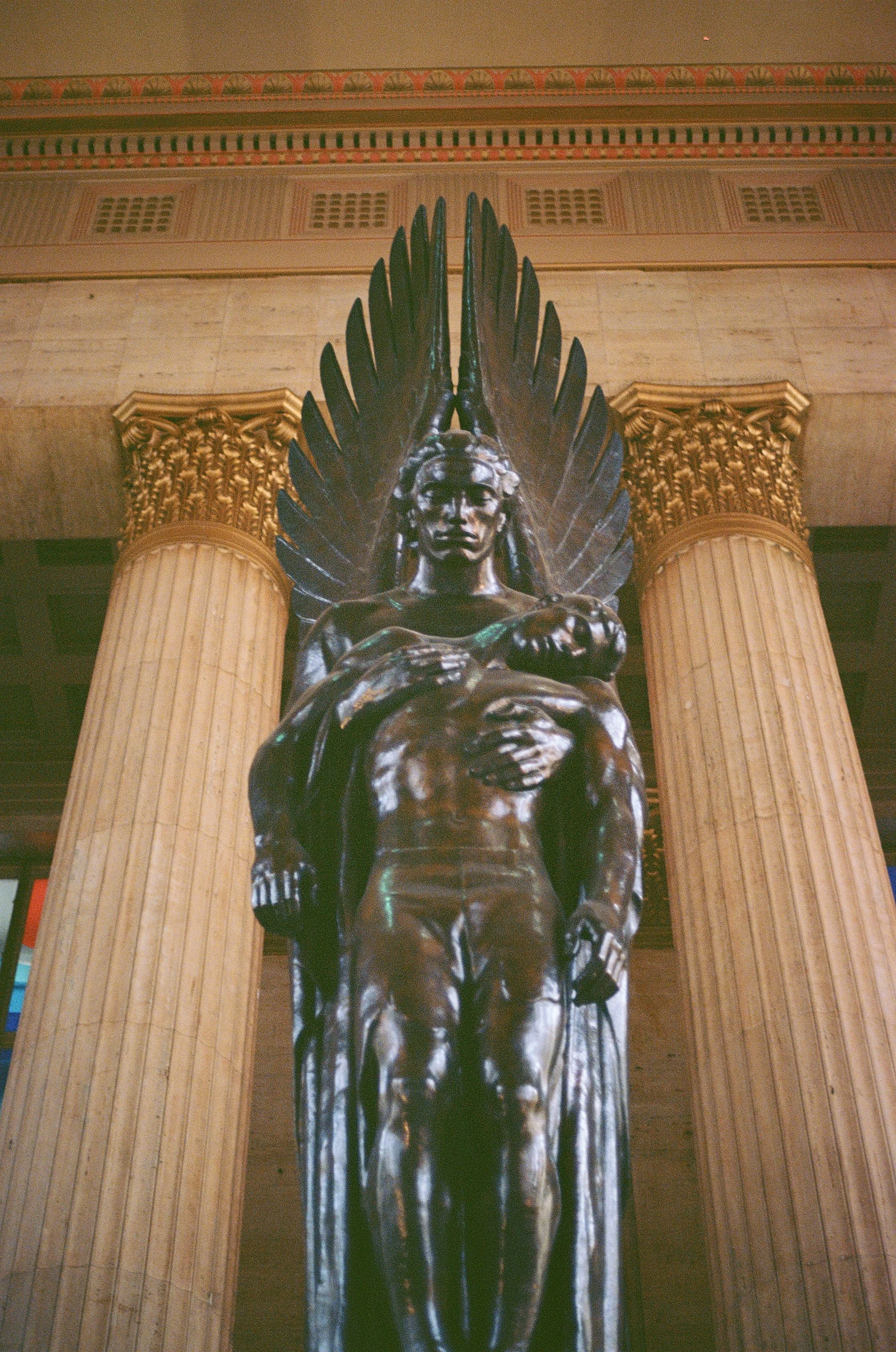A bronze sculpture of a seated woman with large wings behind her, positioned between two tall, classical-style columns with ornate gold capitals inside a building with a decorative frieze and vents on the wall above.
