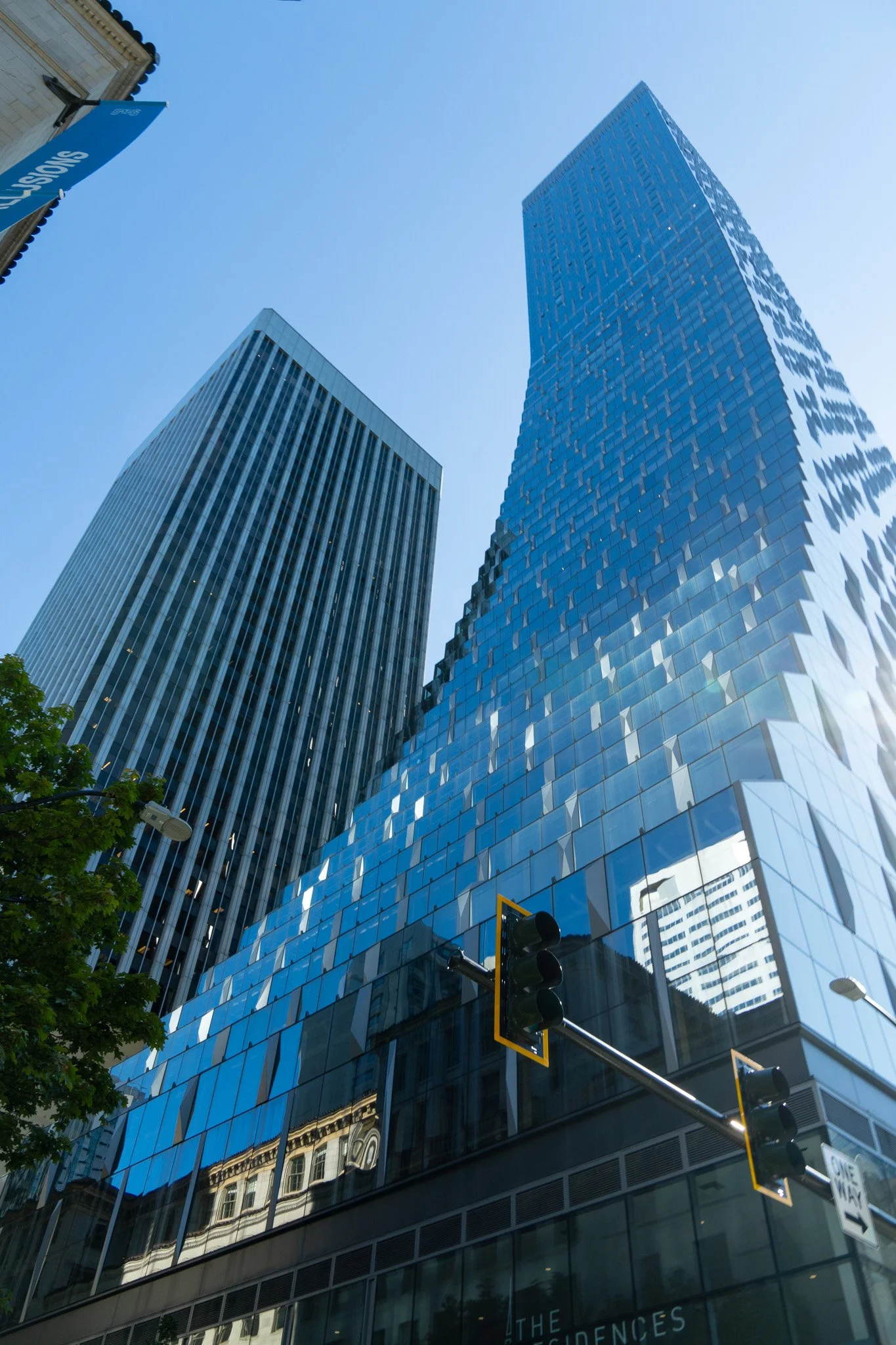 Multiple tall buildings with blue glass facades view from street level during daytime, with traffic signals, trees, and reflections of older buildings in the windows.