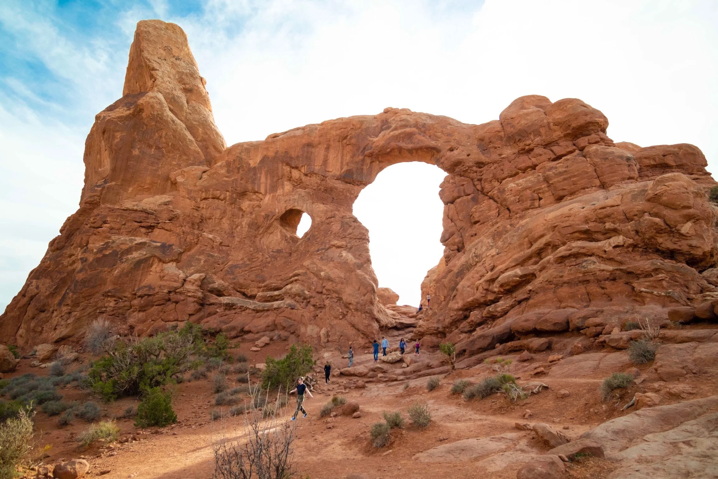 Red rock formation with natural arch in a desert landscape, with people walking on a trail and sparse vegetation.