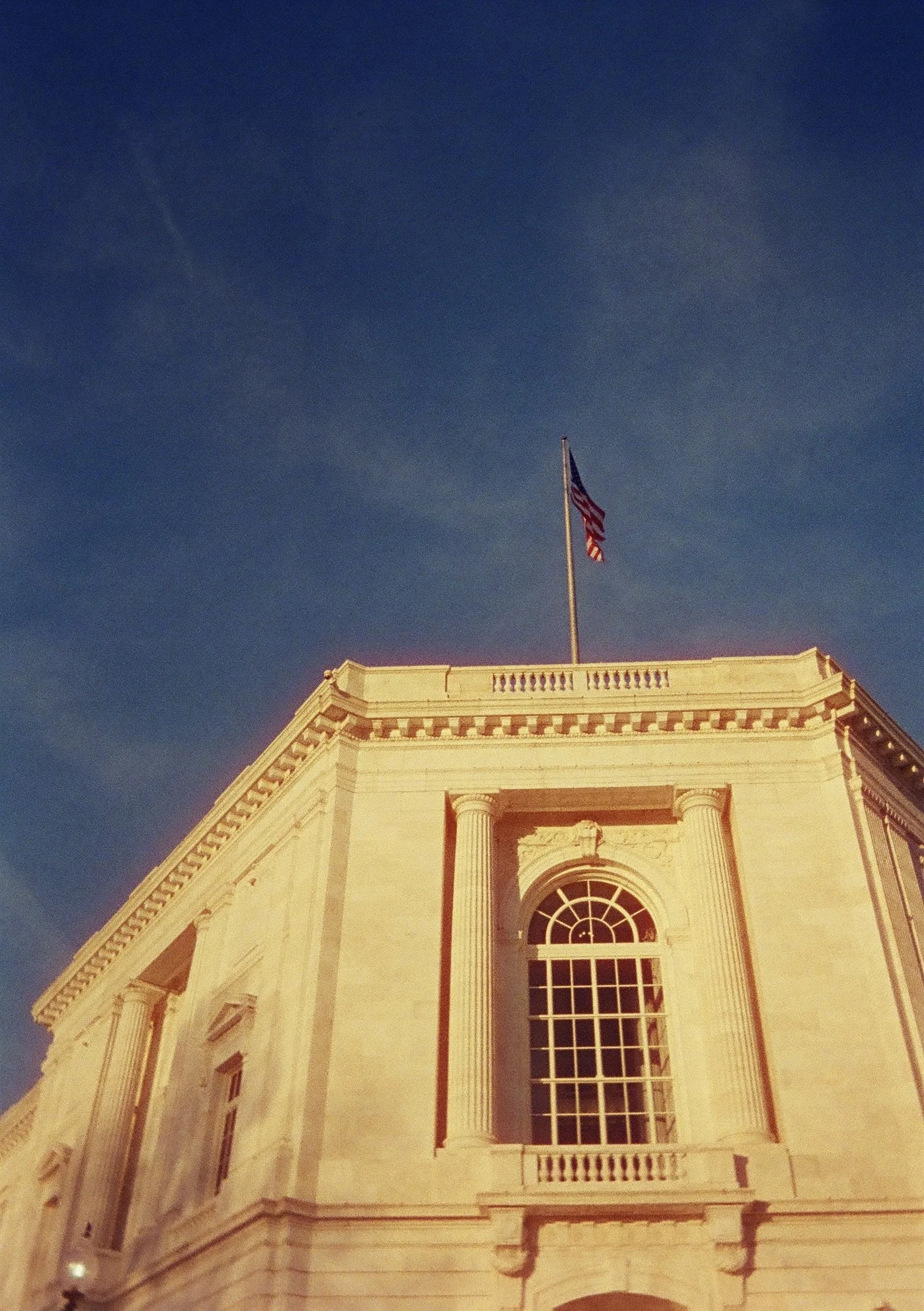 The image shows the top portion of a historic building with classical architecture, featuring large arched windows, columns, and decorative moldings. An American flag is flying on a flagpole on the roof, against a clear evening sky.
