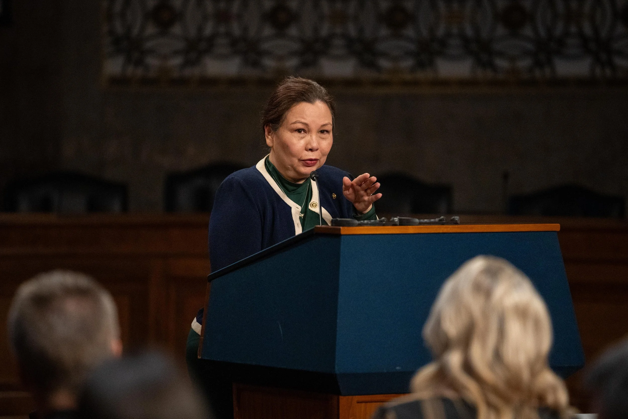 Senator Tammy Duckworth speaking at a podium during a formal event or hearing, with people in the audience listening.