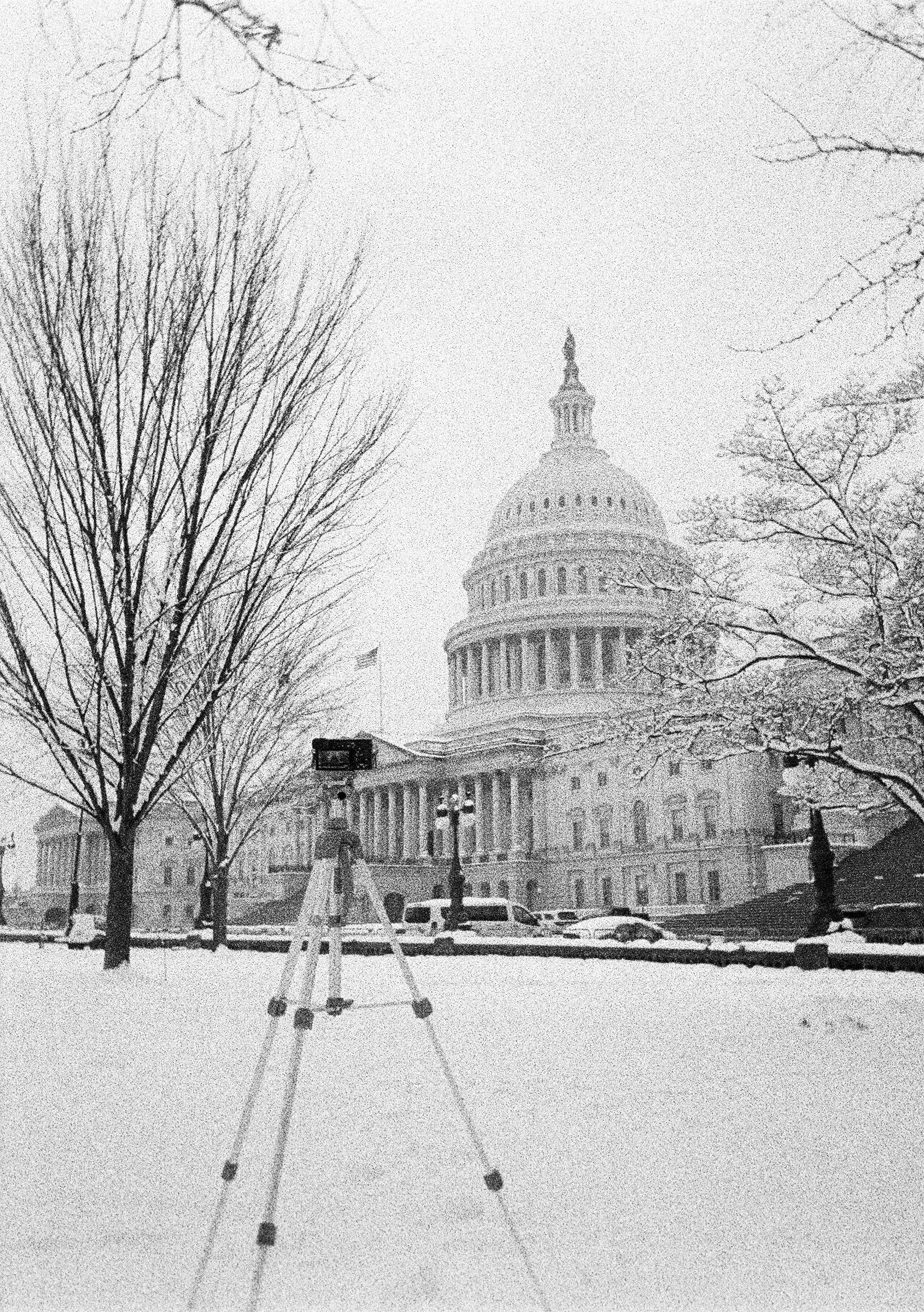 Black and white photo of the U.S. Capitol building with snow on the ground, leafless trees, parked cars, and a snow-covered sidewalk.