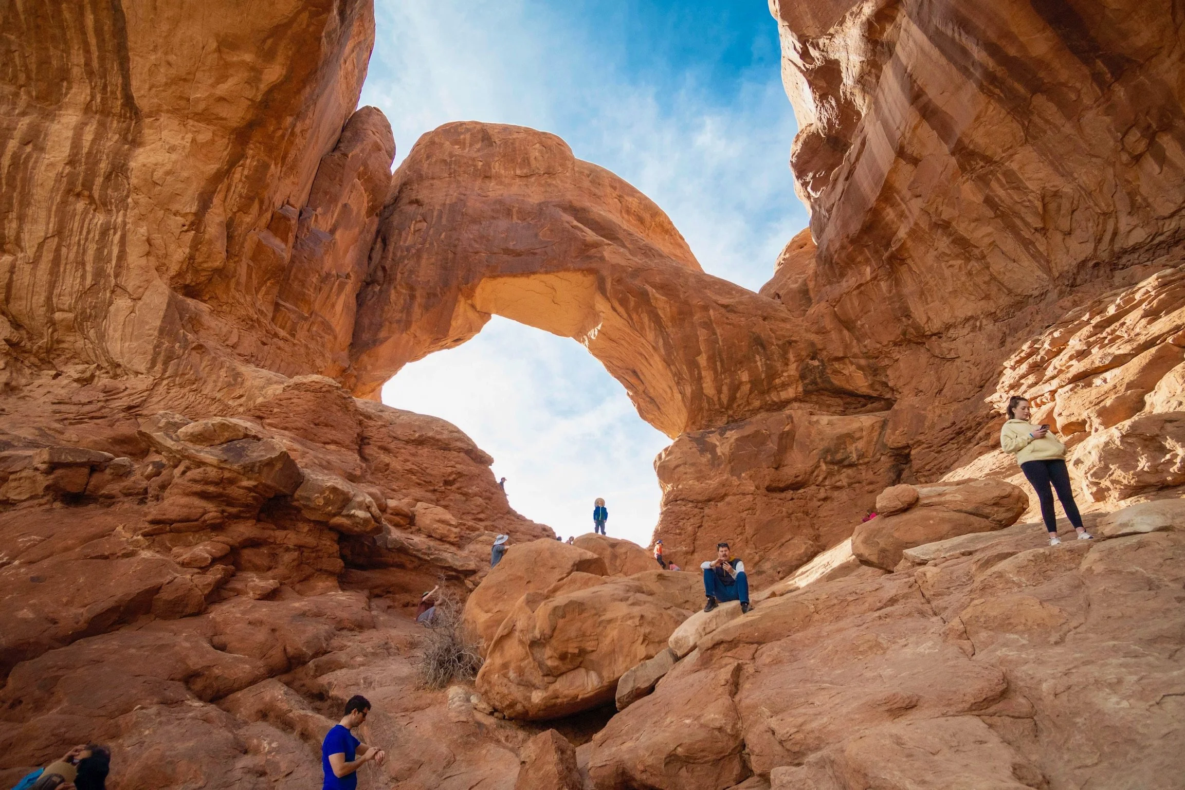 People exploring the red rock formations of a canyon, with a natural arch in the background and a blue sky with some clouds.