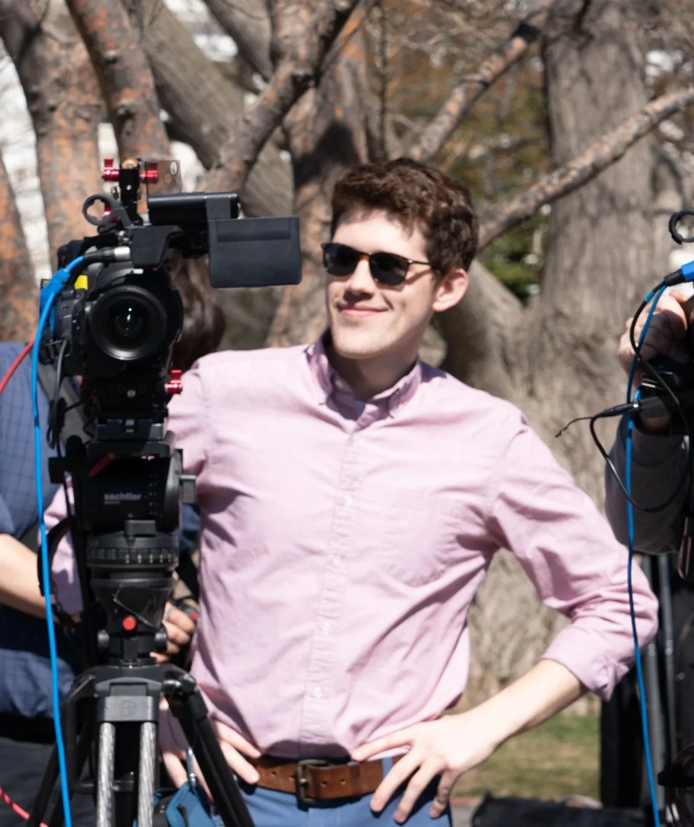 Brooks is standing outdoors in front of a large tree, surrounded by camera equipment, with other people partially visible behind him.