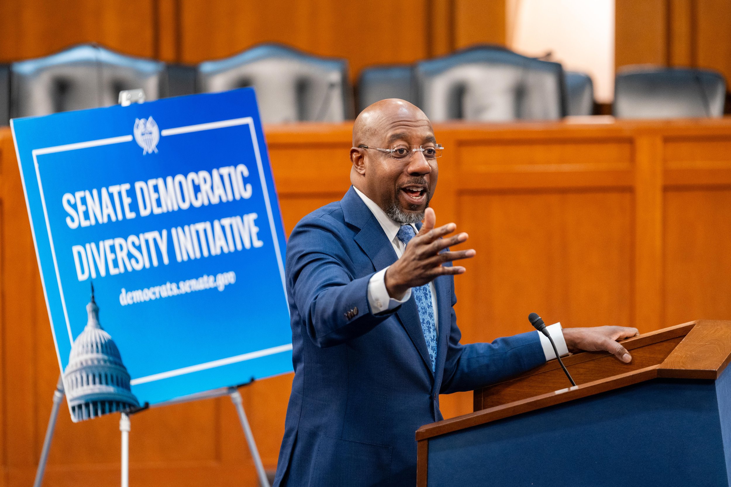 Senator Reverend Raphael Warnock speaking at a podium with a microphone in a hearing room. Behind him is a blue banner that reads 'Senate Democratic Diversity Initiative' and features a graphic of the U.S. Capitol dome.