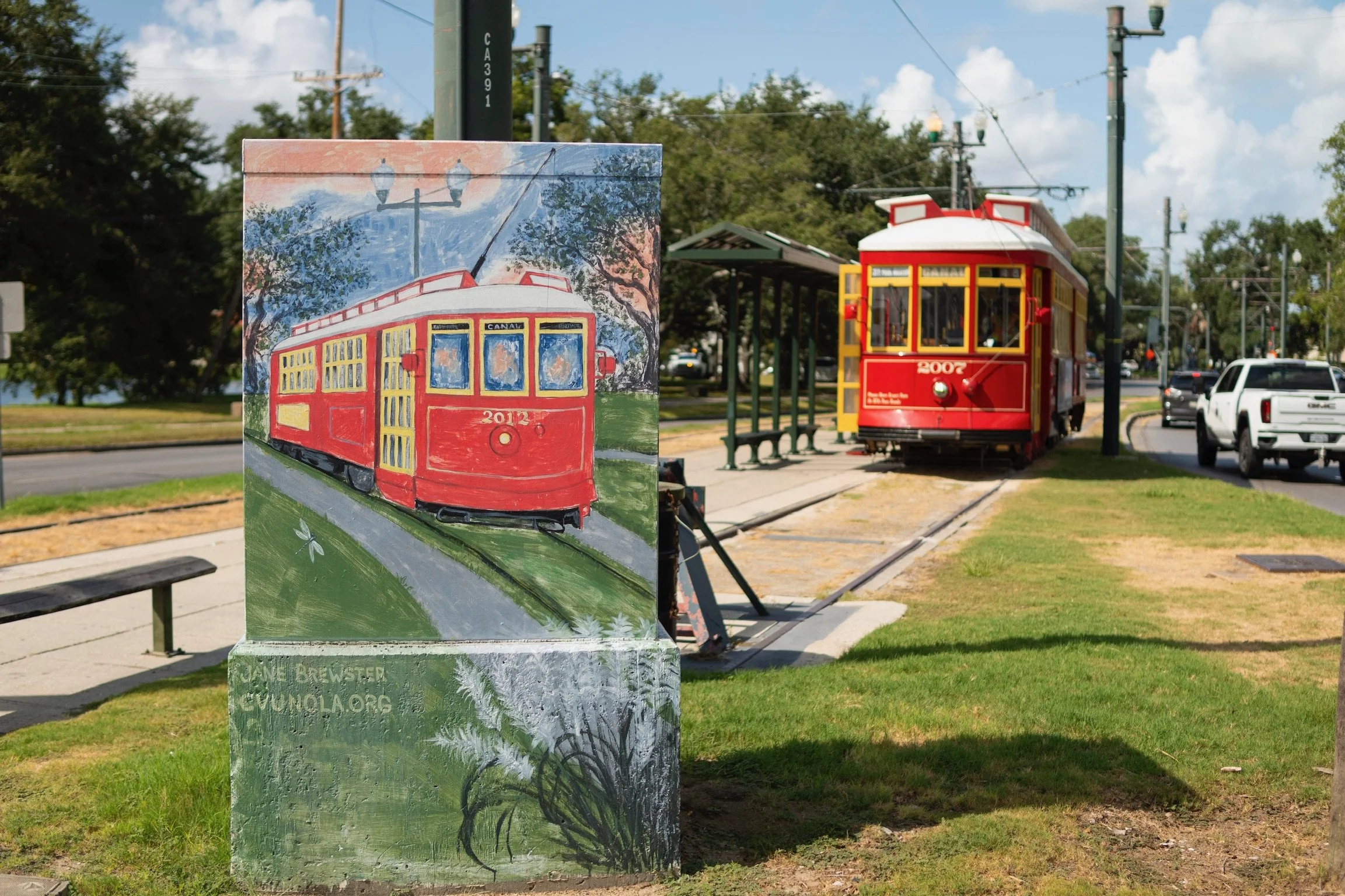 A painted depiction of a red and yellow streetcar with the number 2012 and the word CANAL on the front, displayed on a canvas easel outdoors near a tram stop.