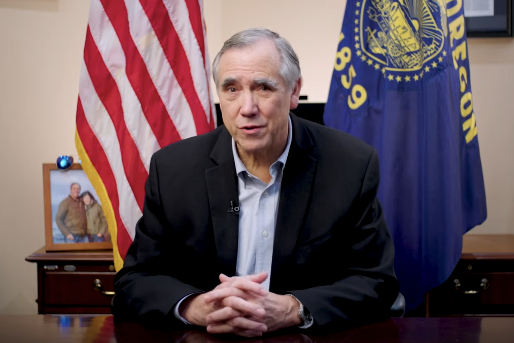 An older man with gray hair in a dark suit and light shirt sitting at a desk, with American flag and another flag in the background, and a photograph on a table behind him.
