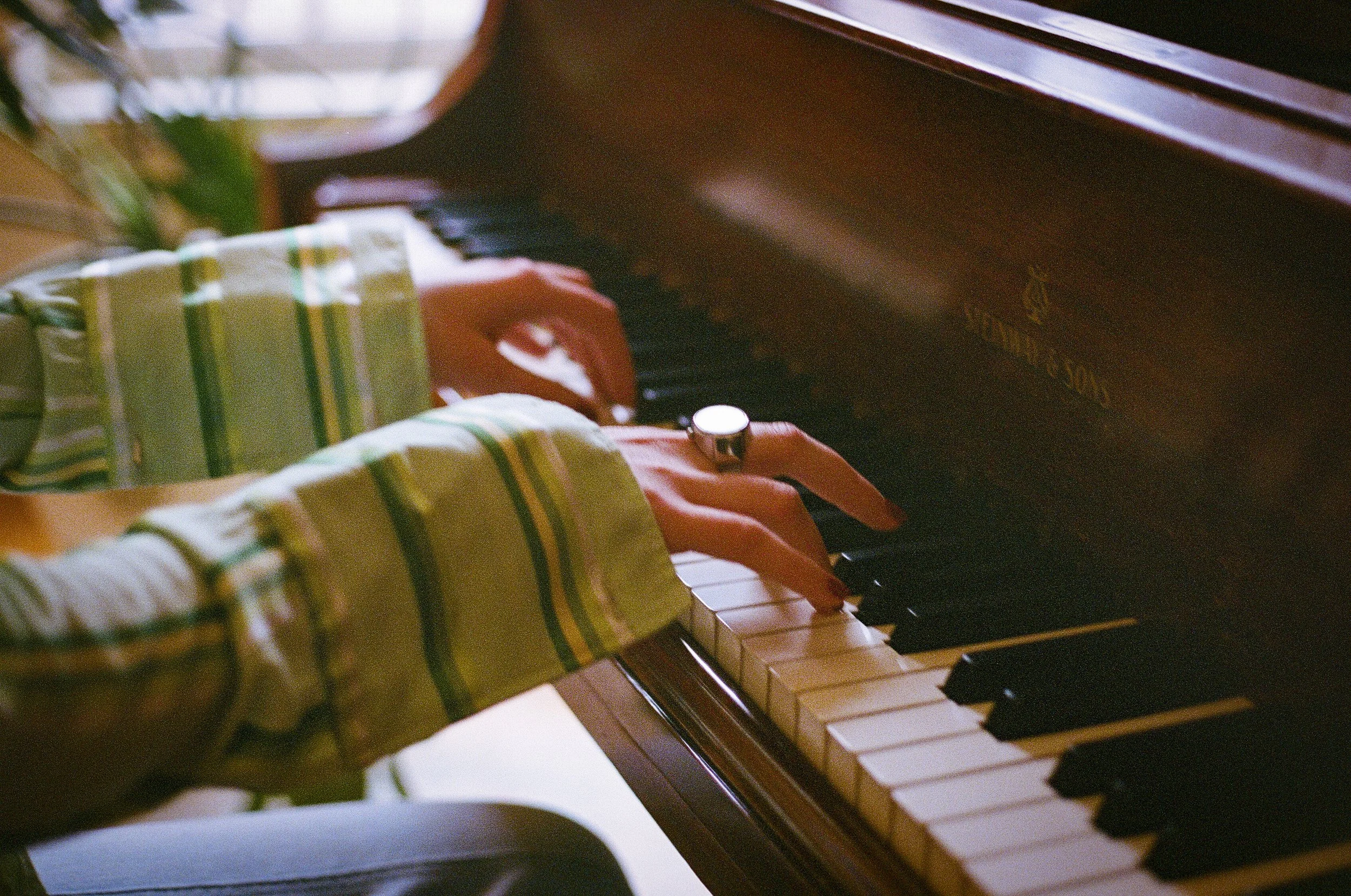 Close-up of a person's hand playing a brown upright piano, with focus on the hand and the keys, wearing a green plaid shirt.