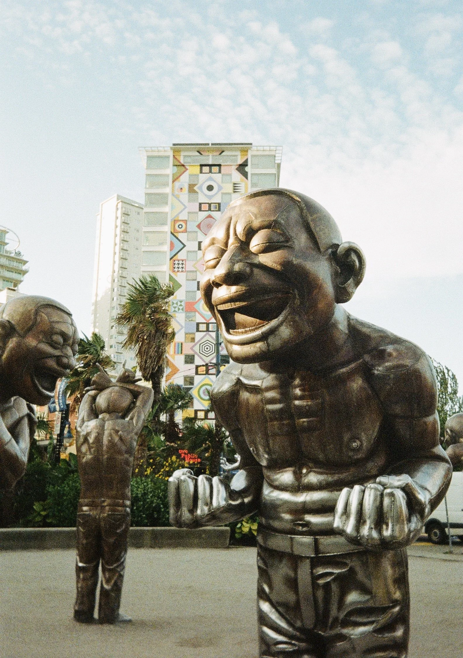 A bronze sculpture of a laughing man in a suit with a tie and striped pants, standing in an outdoor urban area with other similar sculptures and buildings in the background.