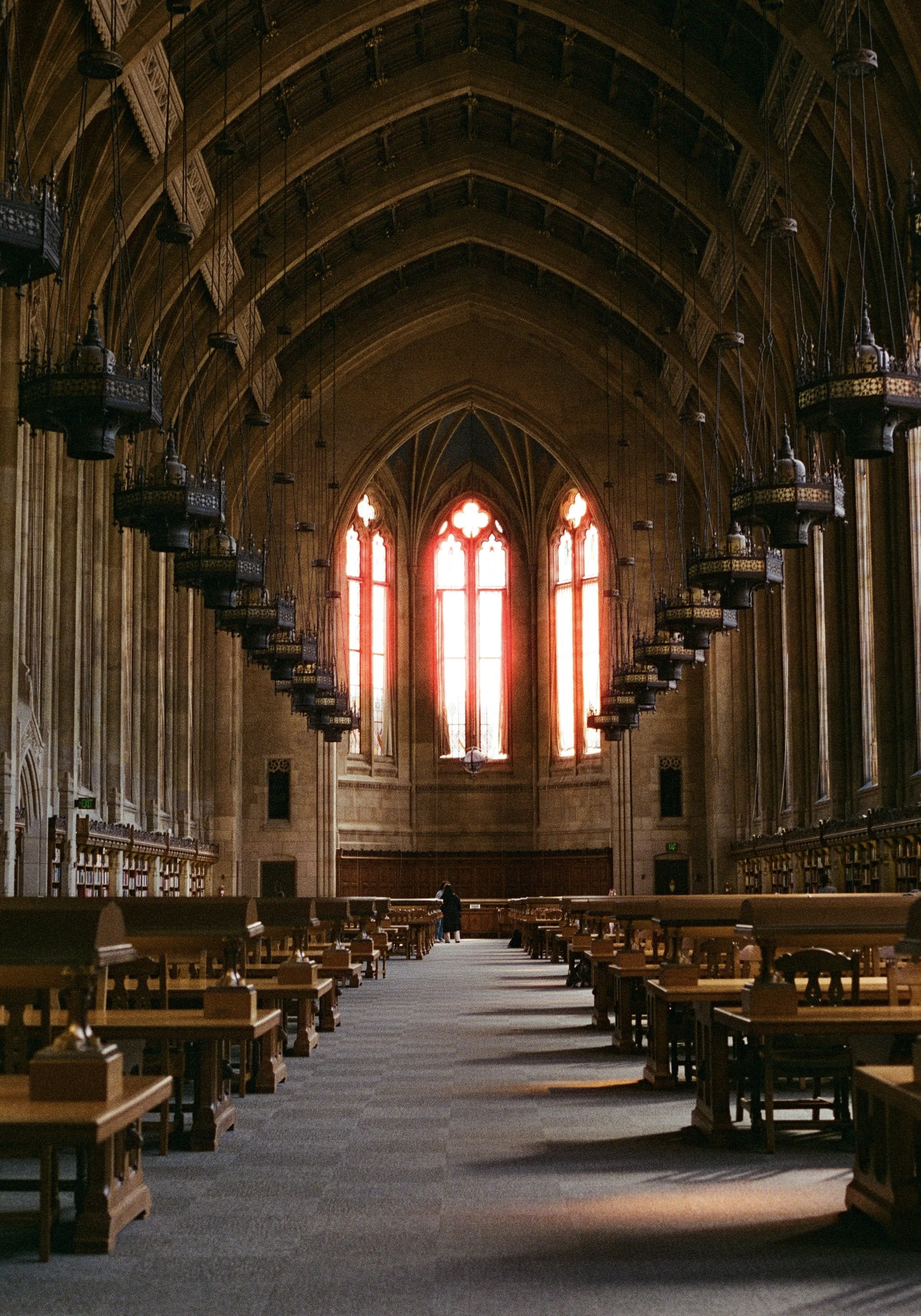 Interior of a large, historic library with high arched ceiling, stained glass windows, hanging lanterns, and rows of wooden tables and chairs.