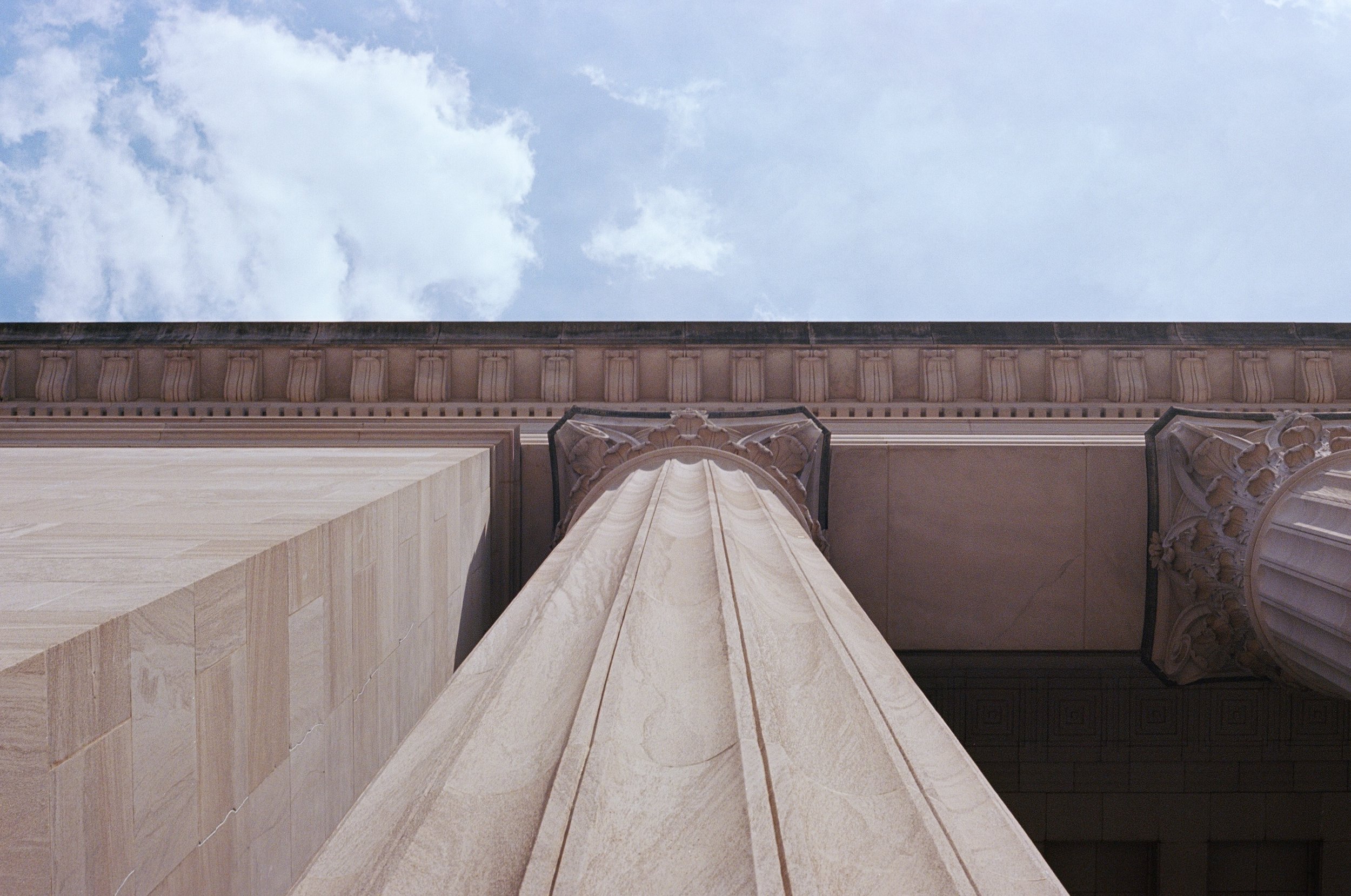 Low-angle view of a stone building facade with decorative columns and a cloudy sky in the background.