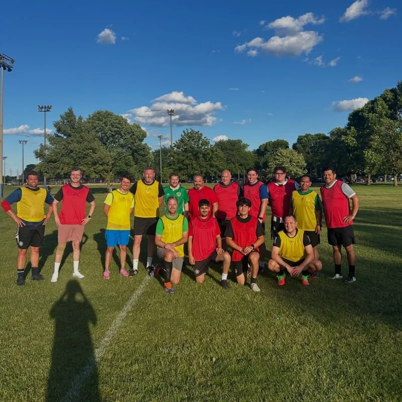 Group of soccer players in colorful jerseys on a grassy field with trees and blue sky in the background.