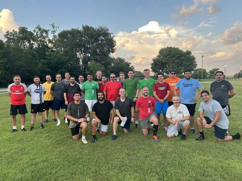 Group of men on a grassy field posing for a photo, some standing and some kneeling, under partly cloudy sky.
