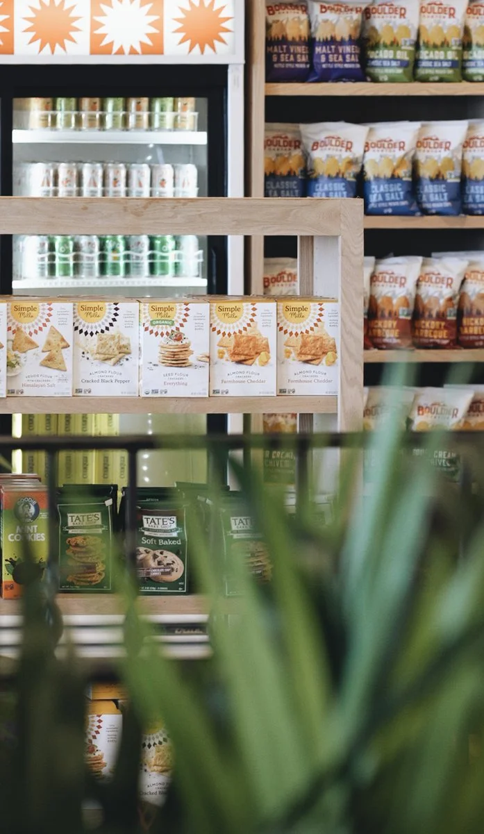Shelves of snack products in a grocery store, including almond flour packets, Boulder sea salts, and cookies, with a view of a refrigerator and stacks of canned drinks in the background.