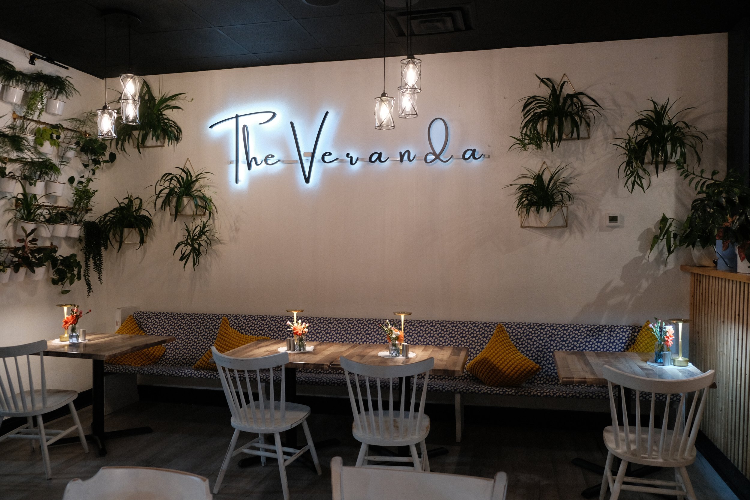 A cozy restaurant interior with a wall decorated with a neon sign that reads 'the Verona.' The wall has several potted plants, and there are tables with chairs and small flower arrangements, creating a warm and inviting atmosphere.