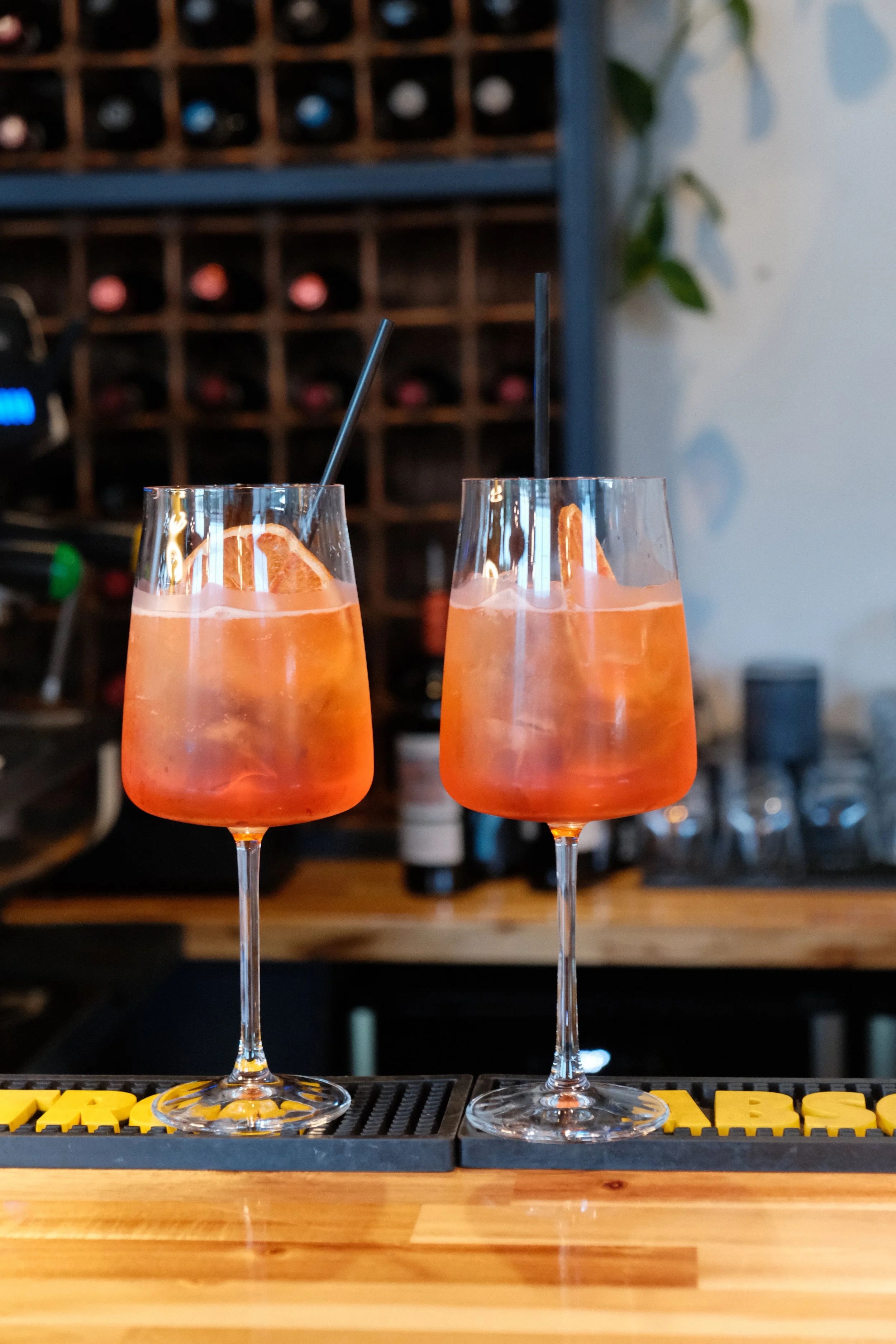 Two tall glasses filled with pinkish-orange cocktail with orange slices and black straws, placed on a bar mat on a wooden bar counter.