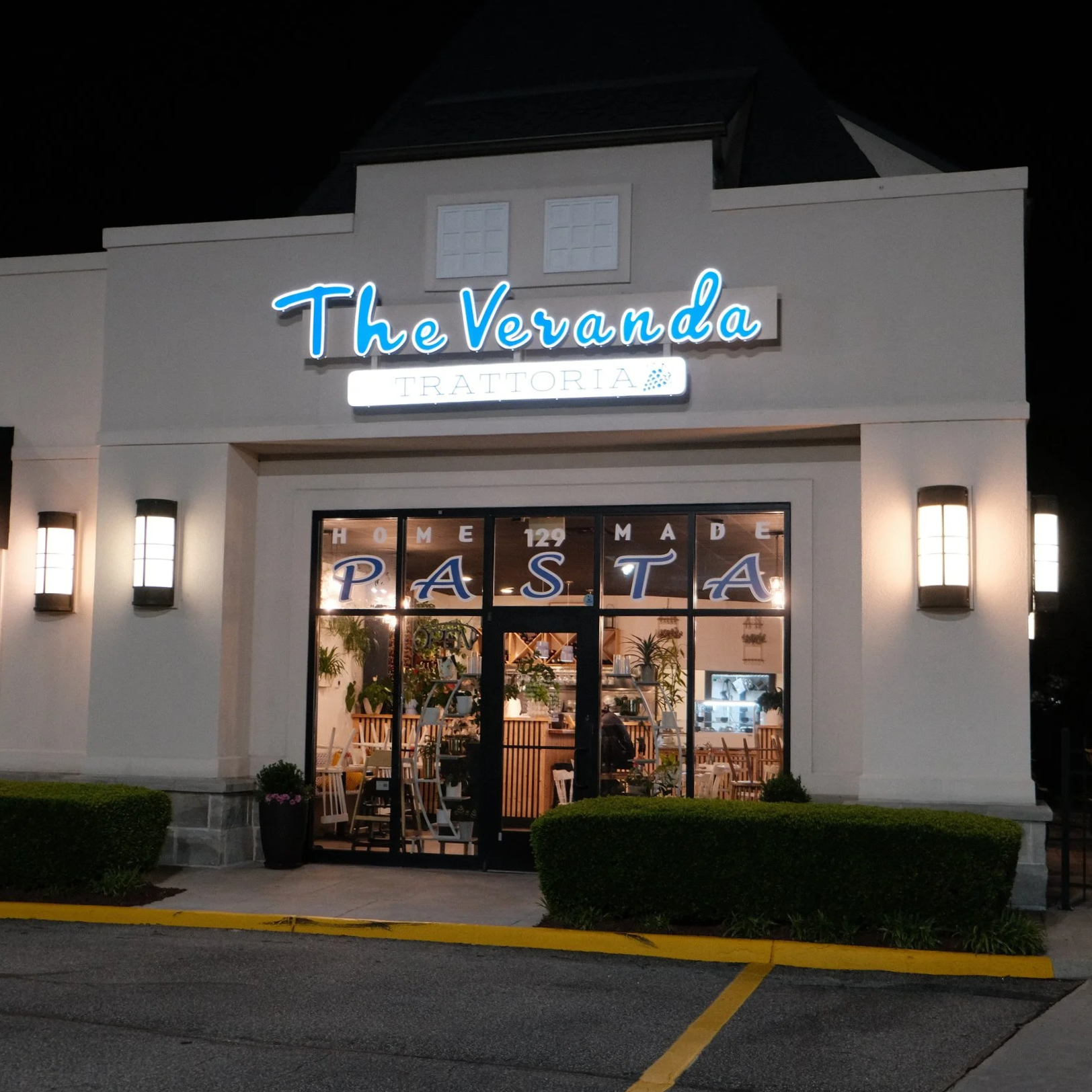 Night view of the exterior of a restaurant called The Veranda Trattoria, with illuminated signage and large glass windows showing the interior dining area with wooden chairs and plants. The building has white walls and is lit by wall-mounted lights. 