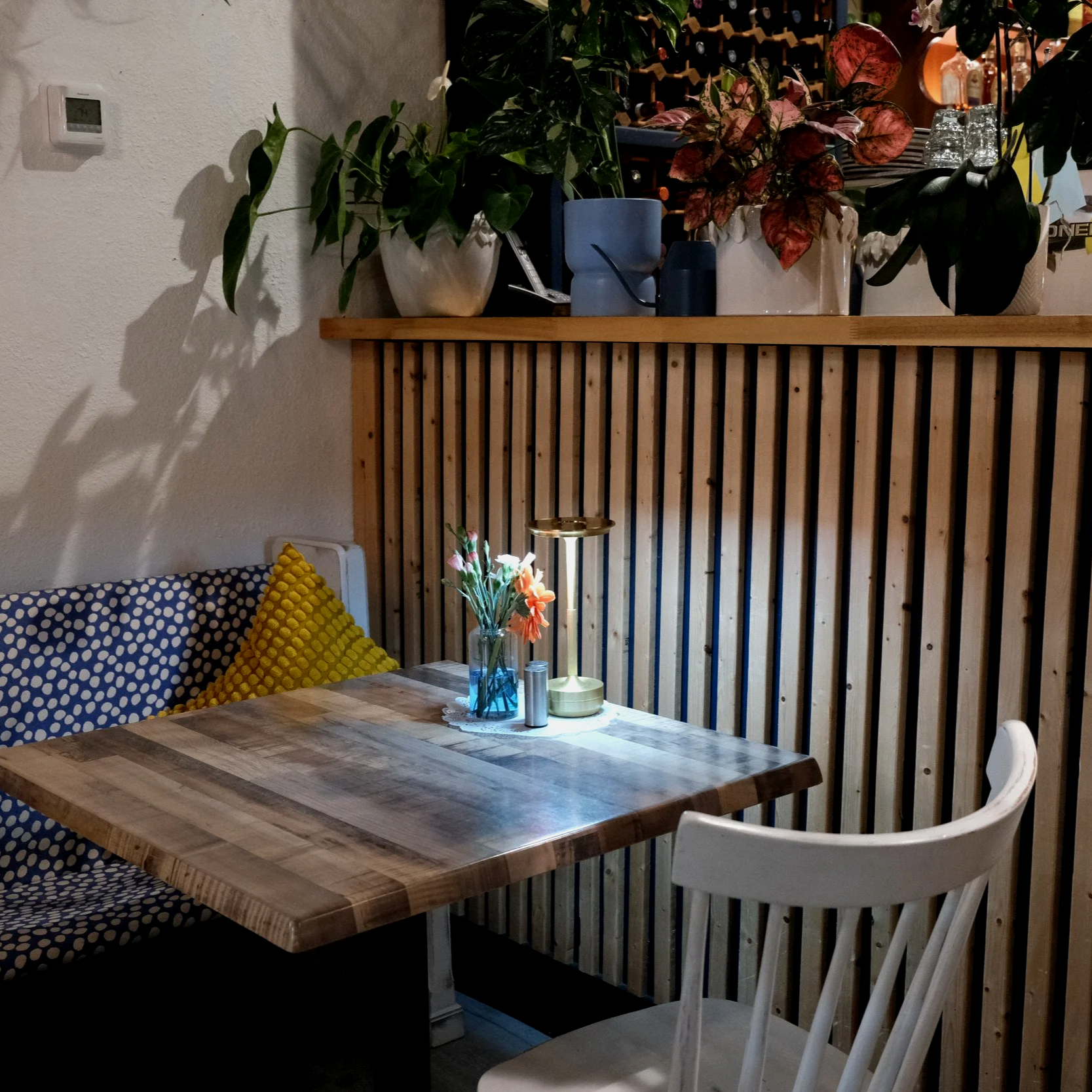 A cozy dining area with a wooden table, white chairs, and yellow textured cushions. The table has a glass vase with flowers, a small candle, and a metallic container. Behind the table, there's a wooden partition with potted plants on top, and a wall 