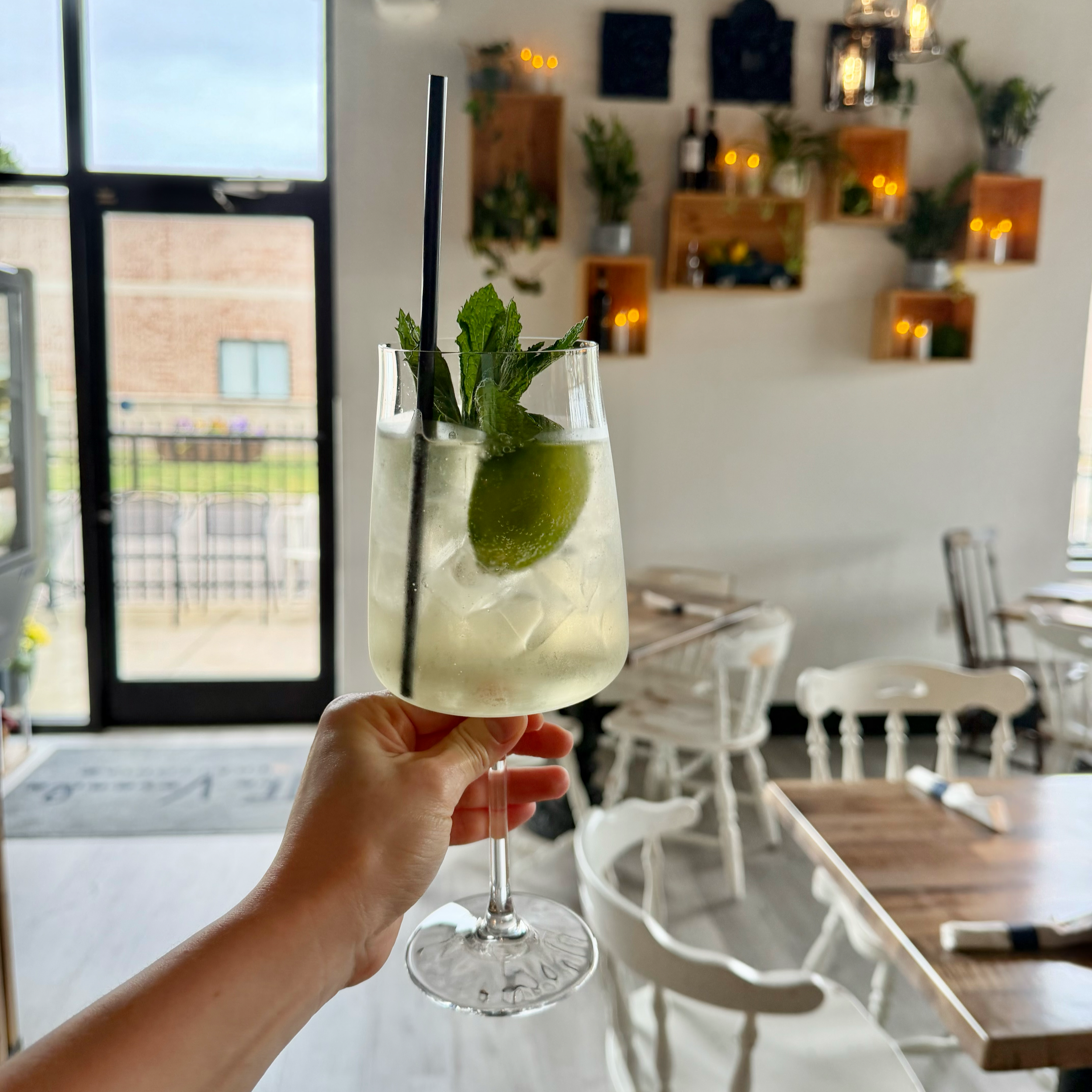 A hand holding a clear glass with a Hugo Spritz garnished with mint leaves  a bright room with large windows. In the background, there are white chairs, a wooden table, and a wall decorated with small wall-mounted planters and lights.