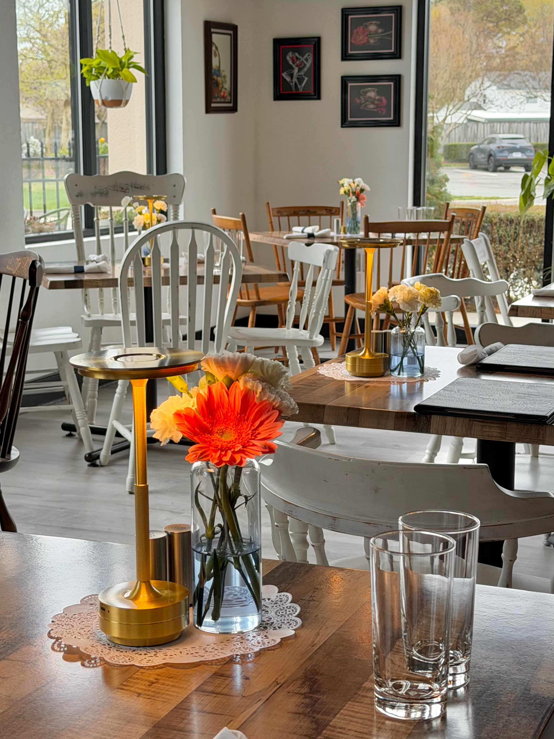 A dining table with flowers, a gold table lamp, and glasses in a well-lit restaurant with artwork on the wall and large windows showing outdoor scenery.