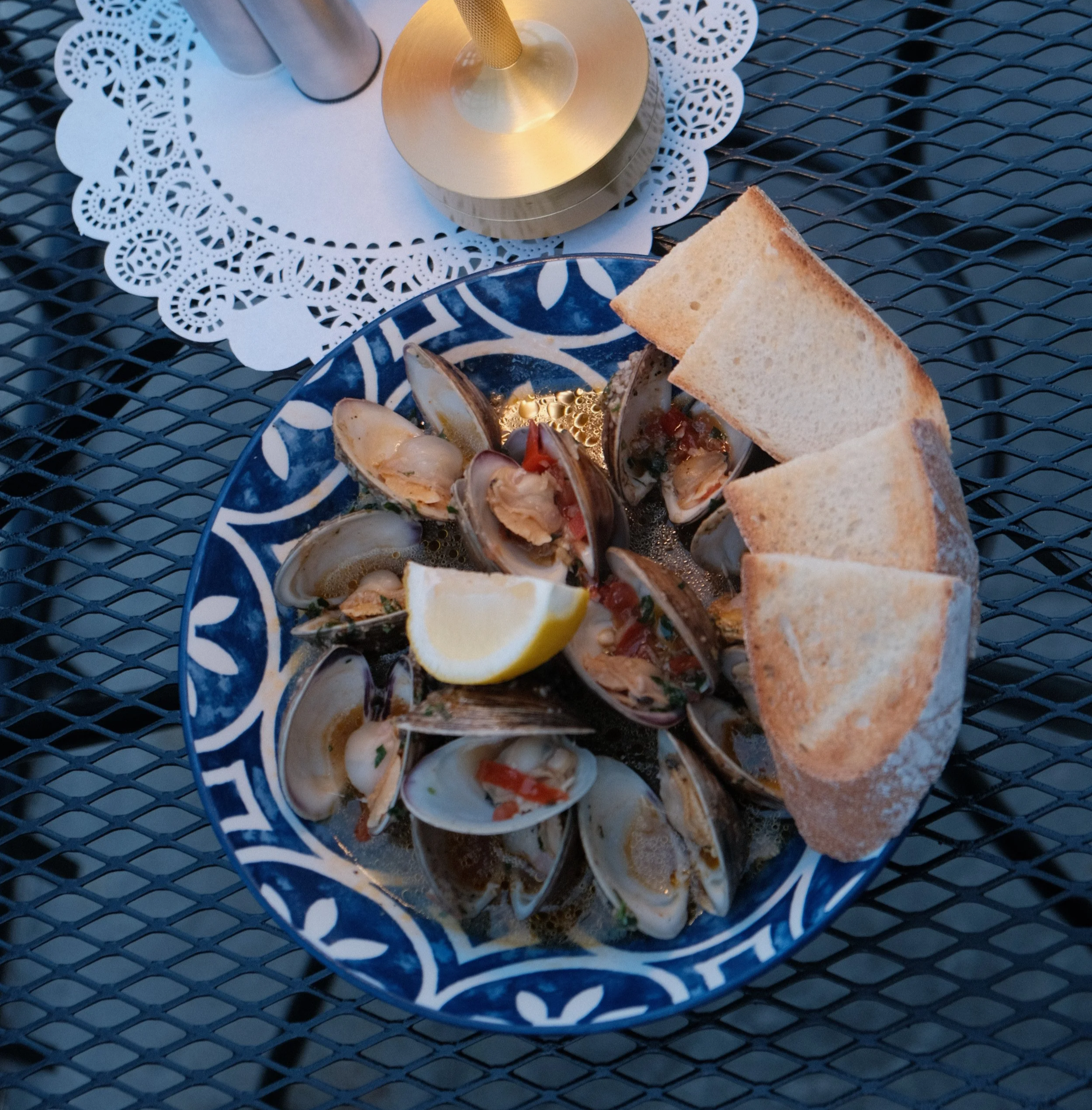A table with a white paper doily, a tall glass with a metallic base, a gold-colored candle holder, a plate with bread and clams, lemon wedges, and a small bowl of seafood with shells.