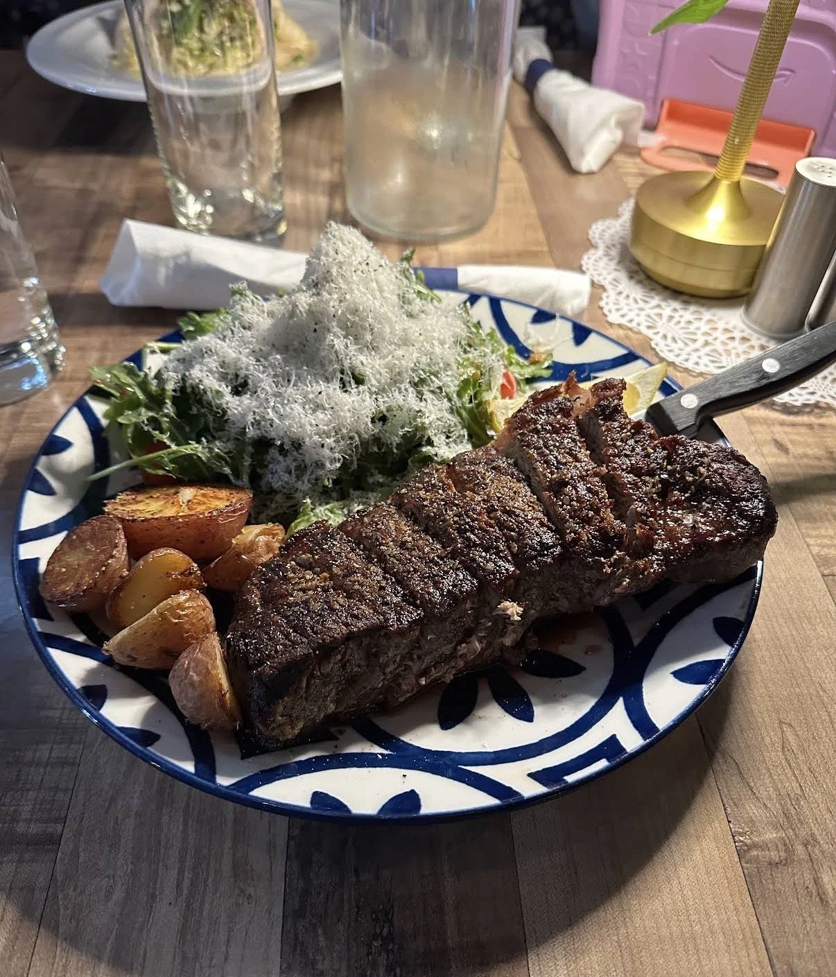 A plate of grilled steak, roasted potatoes, and a salad topped with shredded cheese, on a blue and white patterned dish.