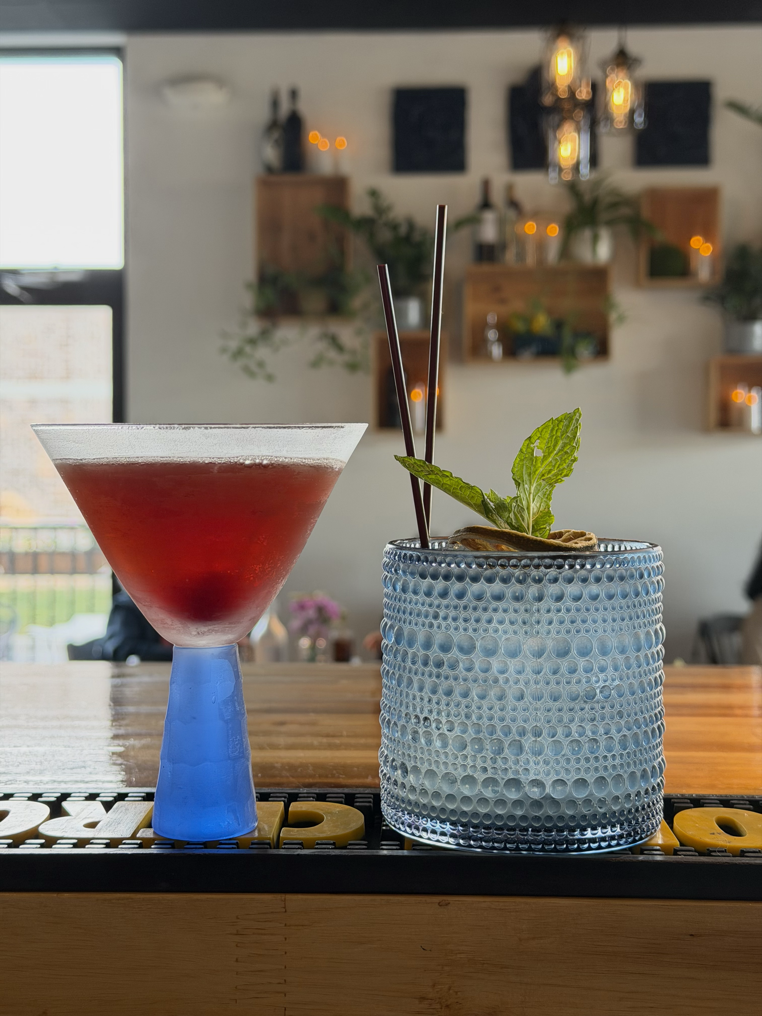 A close-up of a red cocktail in a martini glass with garnish in a textured blue glass container, on a wooden table inside a cozy cafe with plants and decorative lights in the background.