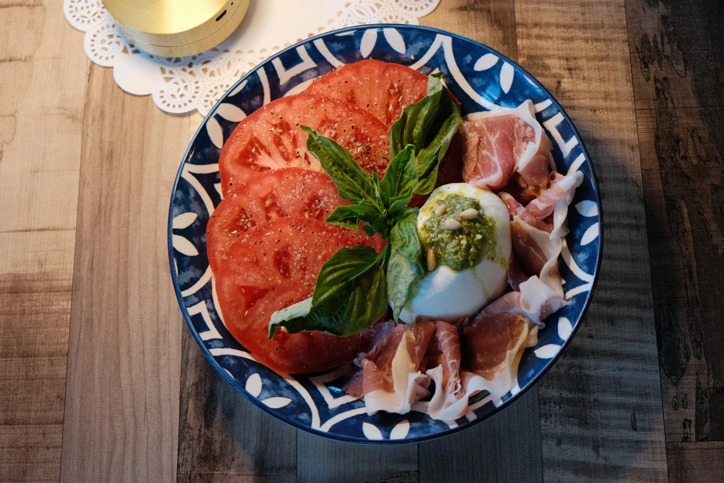 A blue plate with sliced tomatoes, fresh basil, mozzarella cheese topped with pesto, and prosciutto on a wooden table.