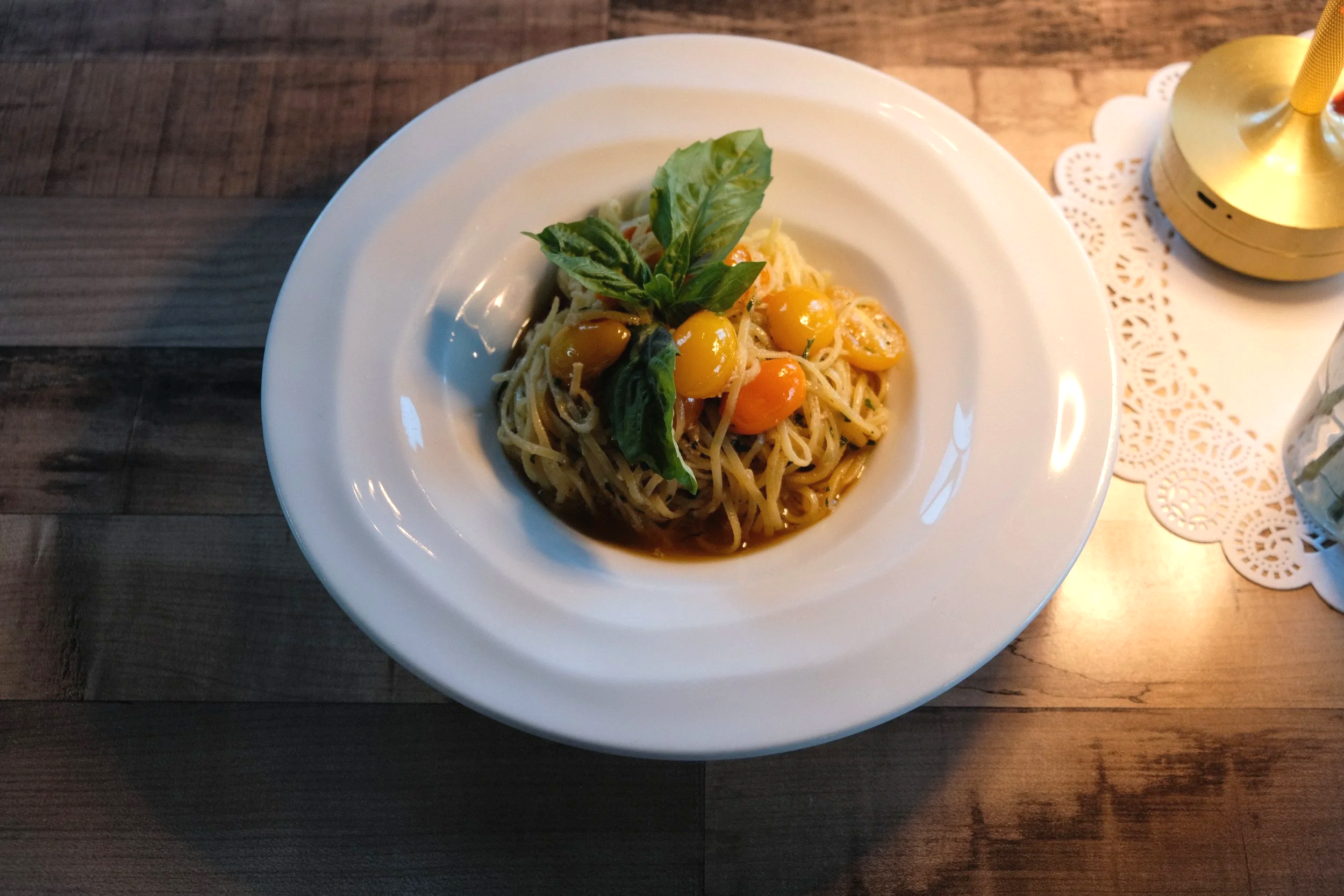 A plate of tagliollini agilo e olio pasta garnished with fresh basil leaves and yellow cherry tomatoes, served on a white dish, with a golden table lamp in the background on a wooden table.
