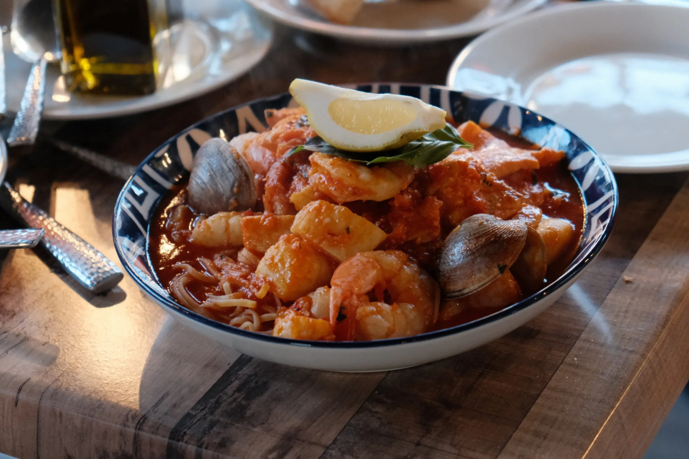 A bowl of seafood stew with shrimp, clams, and fish in a tomato-based broth, garnished with a lemon wedge and fresh herbs, sitting on a wooden table.