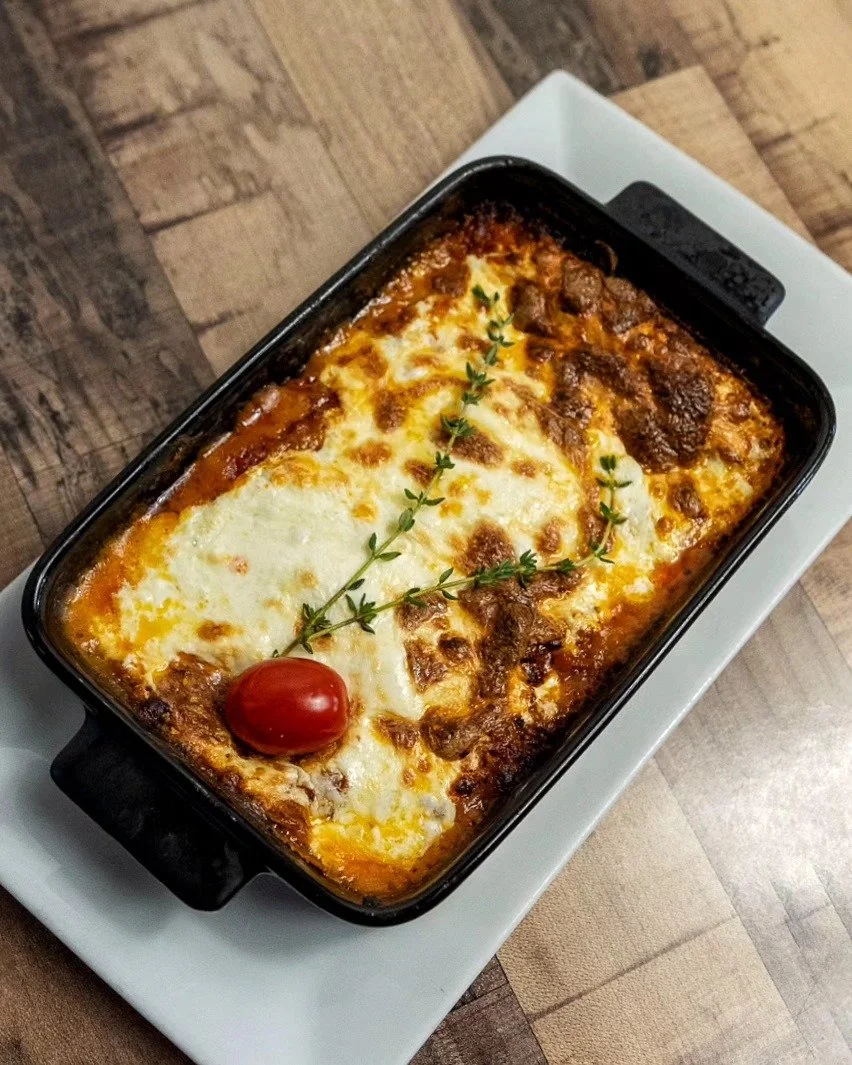 Baked lasagna with melted cheese, cherry tomato, and sprig of herbs in a black baking dish on a white plate.