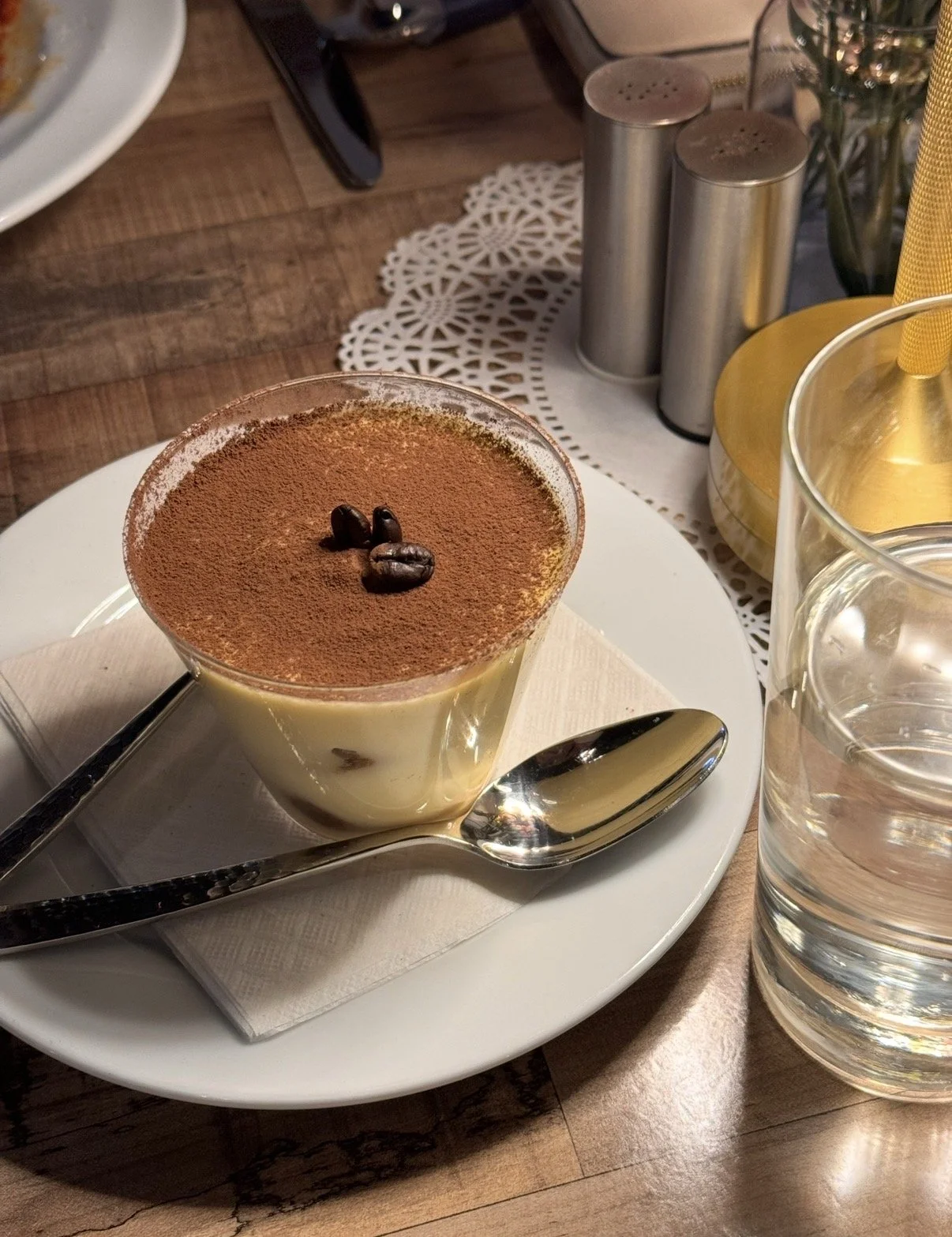 A cup of tiramisu topped with cocoa powder and three coffee beans, placed on a white plate with a small silver spoon and a napkin. Next to it is a glass of water, and in the background, salt and pepper shakers and other table settings are visible.