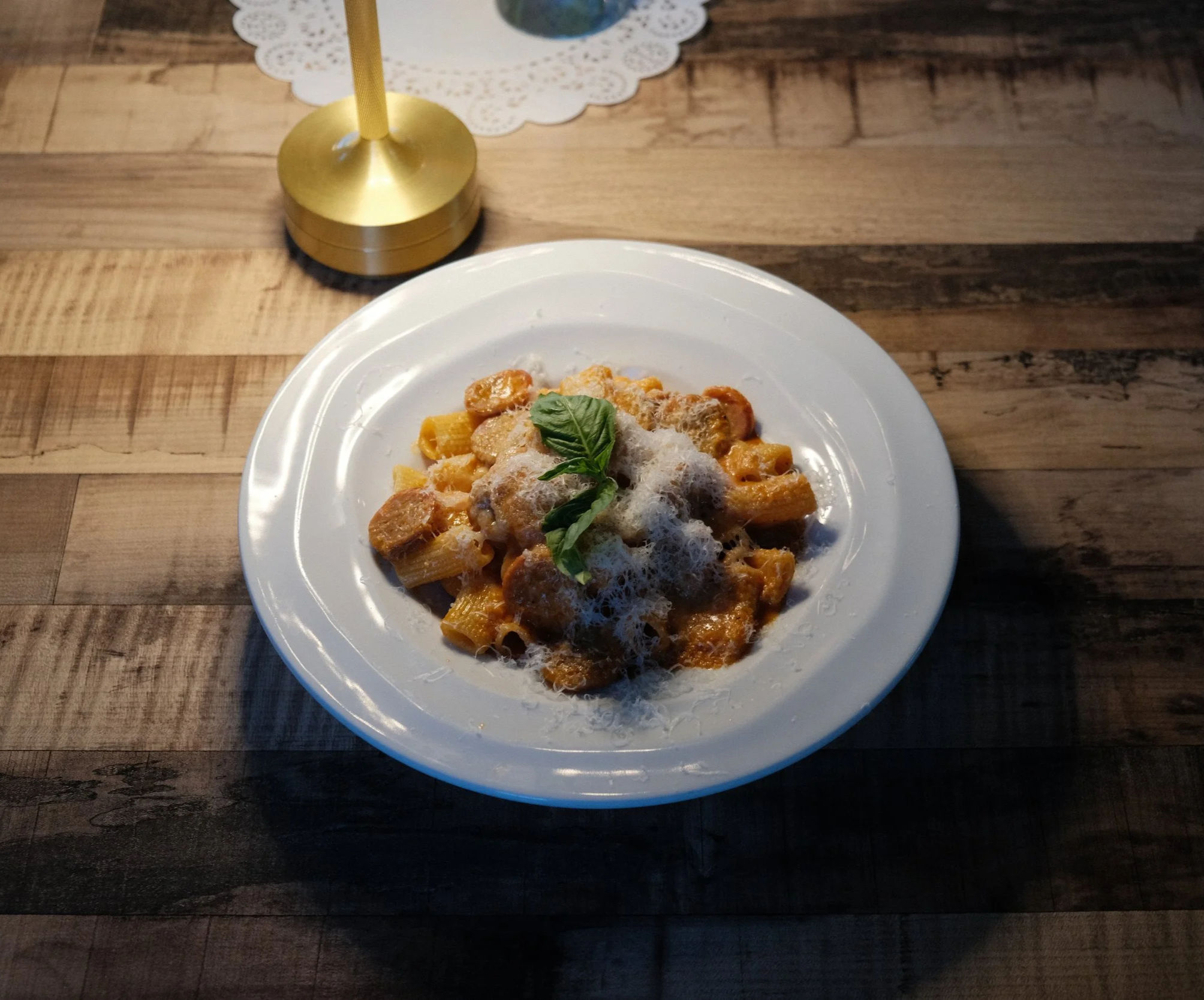 A plate of pasta with vodka sauce, topped with cheese and basil, on a wooden table. A gold-colored table lamp  in the background.