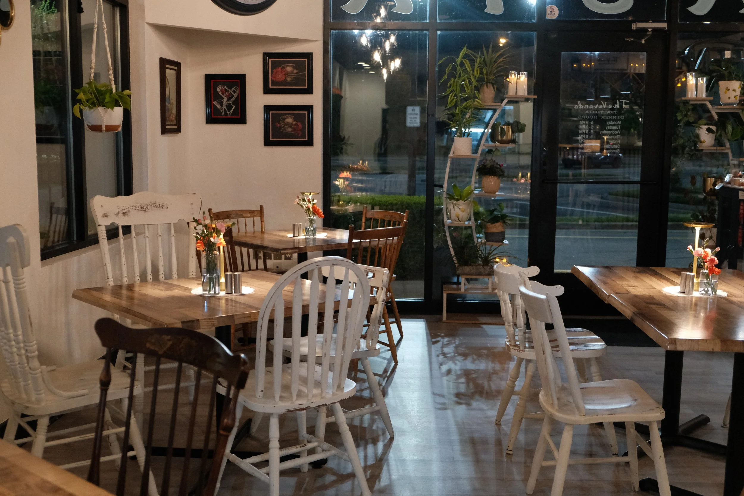 Interior of a cozy cafe or restaurant with wooden tables and white chairs, decorated with flowers, plants, and framed artwork on the walls. Large windows reveal a nighttime street view outside.