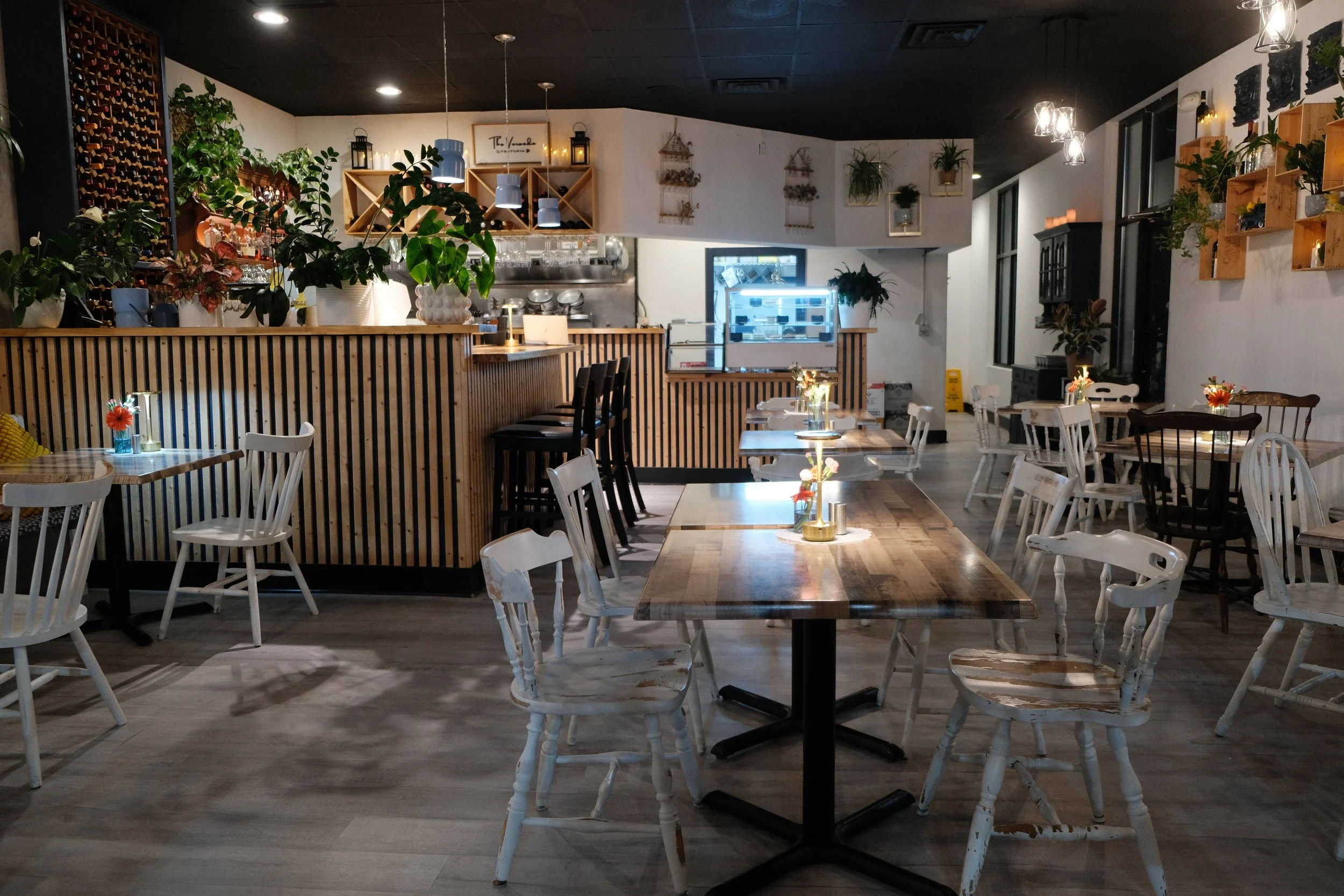 Interior of a cozy restaurant with wooden tables and white chairs, decorated with small vases of flowers. There's a bar area with plants and hanging lights, with shelves of bottles and glassware behind it. The lighting is warm and inviting with a mix