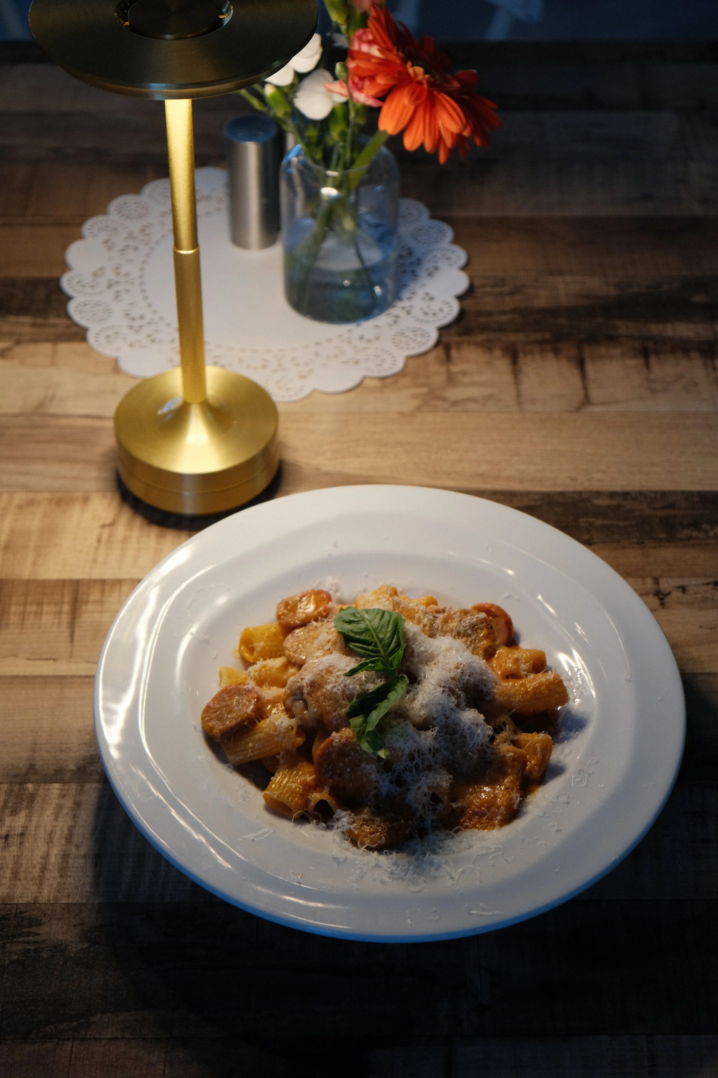 A plate of pasta with tomato sauce, topped with grated cheese and a basil leaf, on a wooden table. In the background, there is a table center with a glass vase of red and white flowers, a white paper doily, a golden candle holder, and a salt shaker.