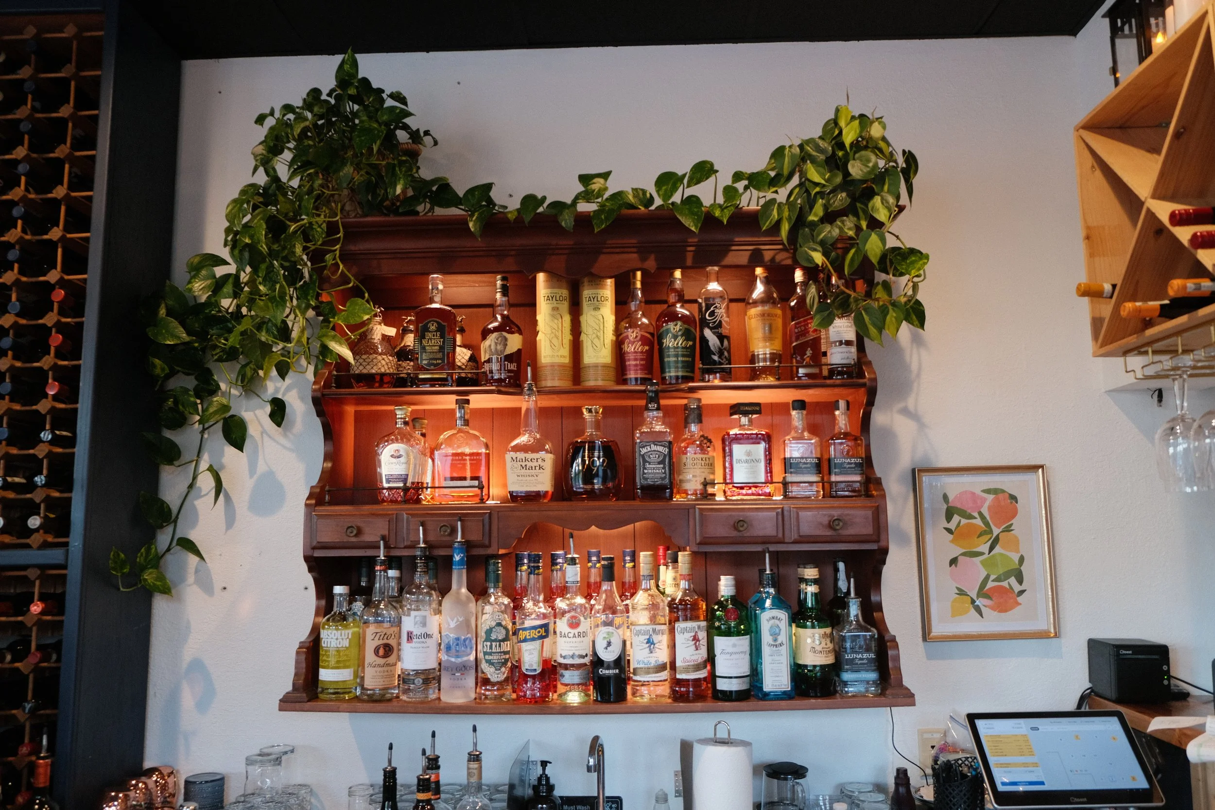 Bar area with shelves of various whiskey bottles and green potted plants, framed art, and a wine rack on the wall.