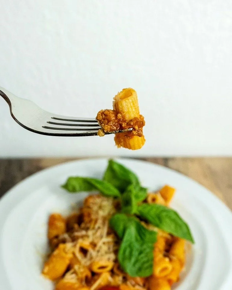 Close-up of a fork holding a small piece of cooked pasta with tomato sauce, above a plate of pasta with basil leaves.