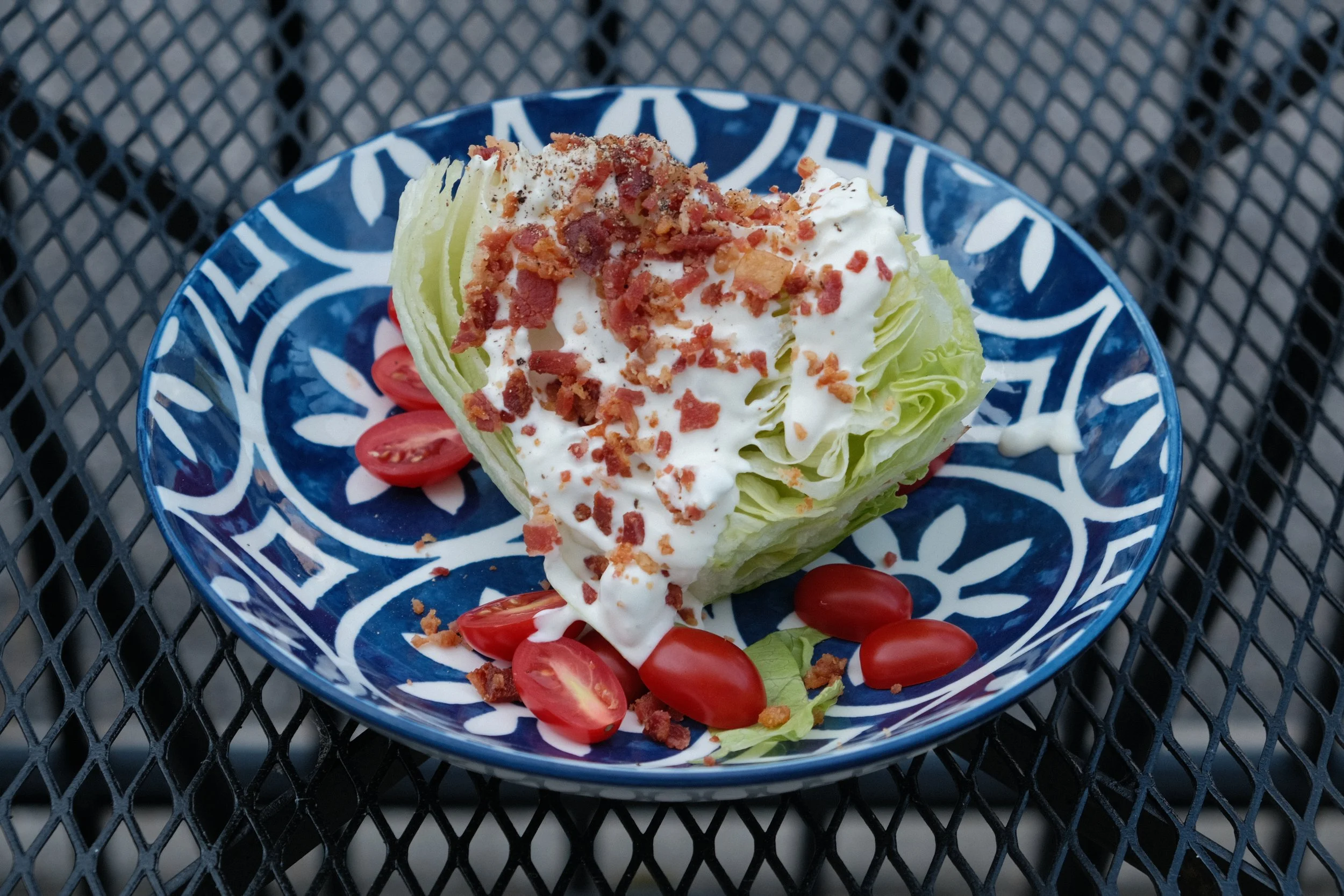 A lettuce wedge topped with creamy dressing, crumbled bacon, and chopped tomatoes on a patterned blue plate, with cherry tomatoes and lettuce leaves around it, on a black metal outdoor table.